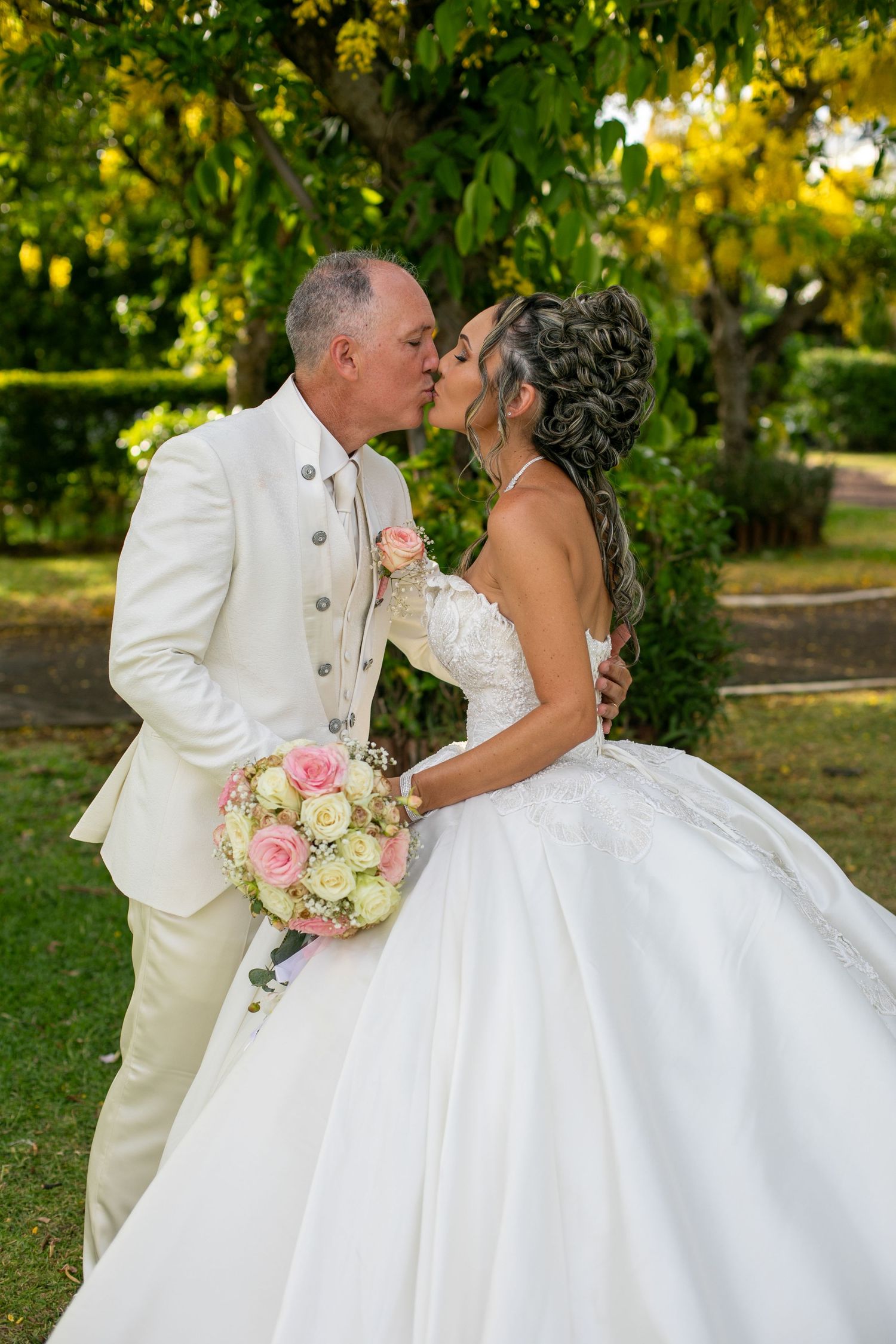 A romantic wedding kiss between a couple in white attire under lush green trees.
