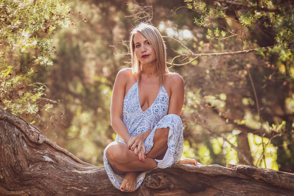 Person in light blue dress sitting on tree branch in sunlit forest setting.
