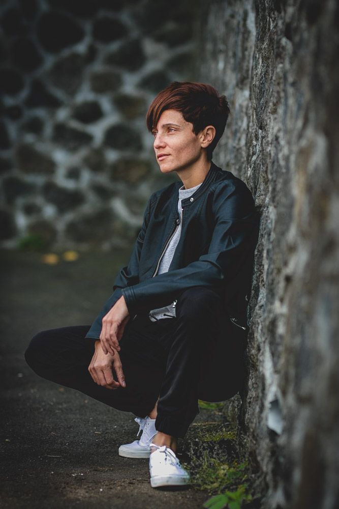 Person in black jacket sitting against stone wall during outdoor photoshoot.