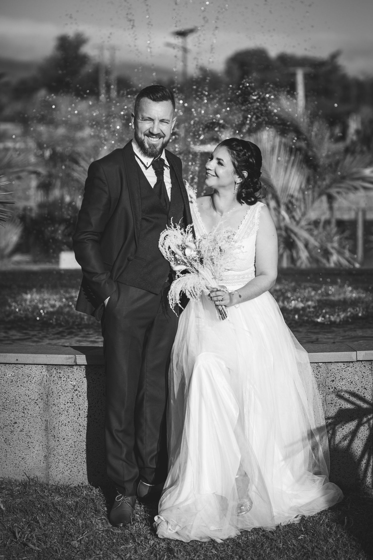 A wedding couple shares a joyful moment together in front of a fountain in black and white photography.