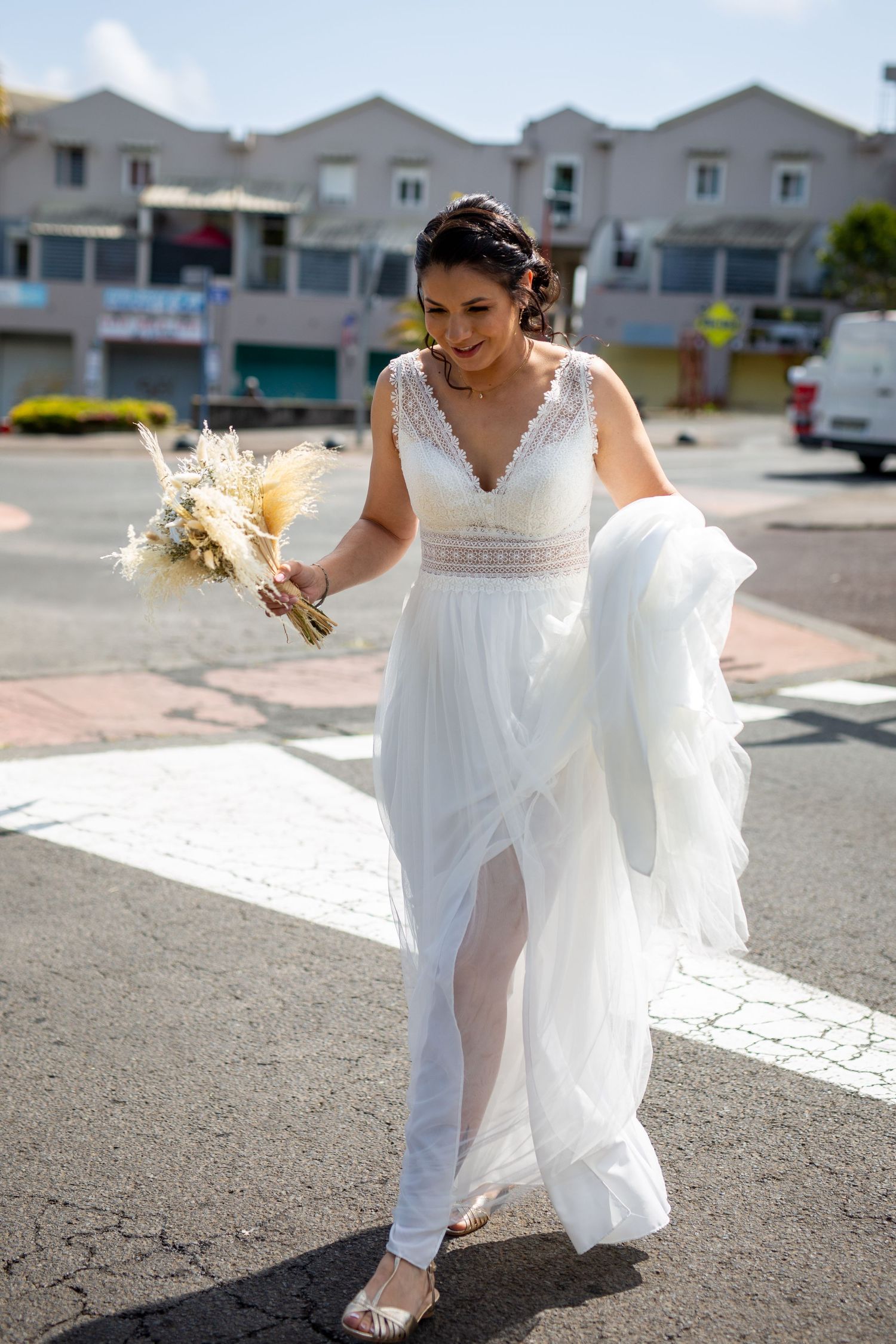 A bride in a flowing white dress with lace details holds a dried floral bouquet while walking on a sunny day.