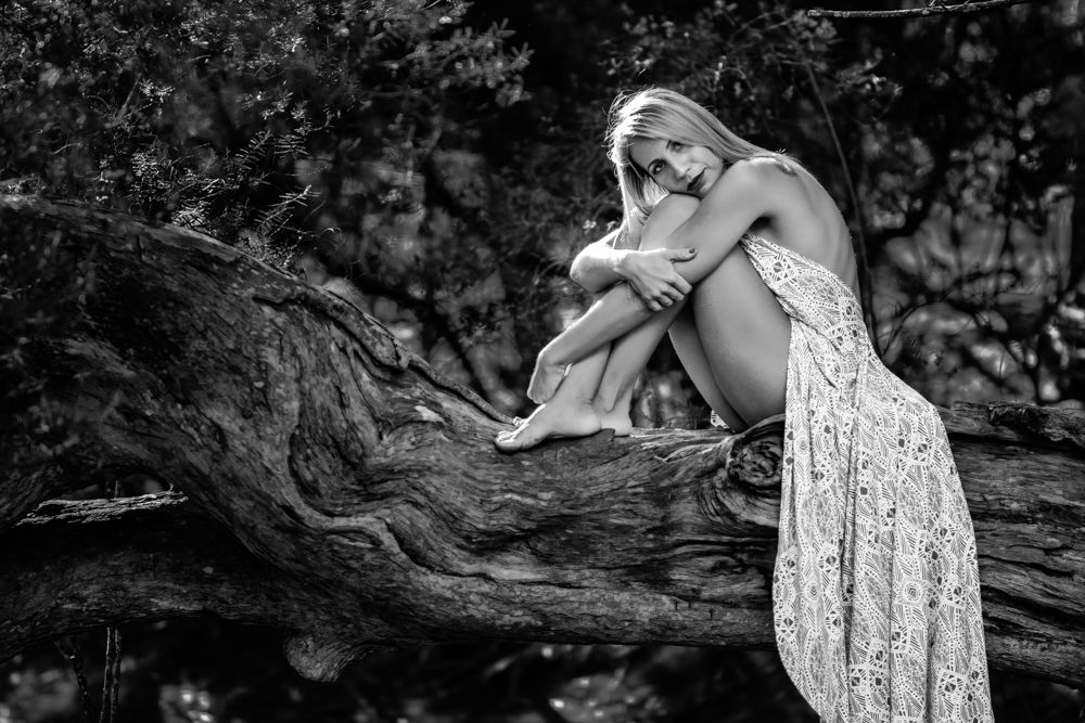 Black and white portrait of a figure in flowy dress resting on a tree branch.