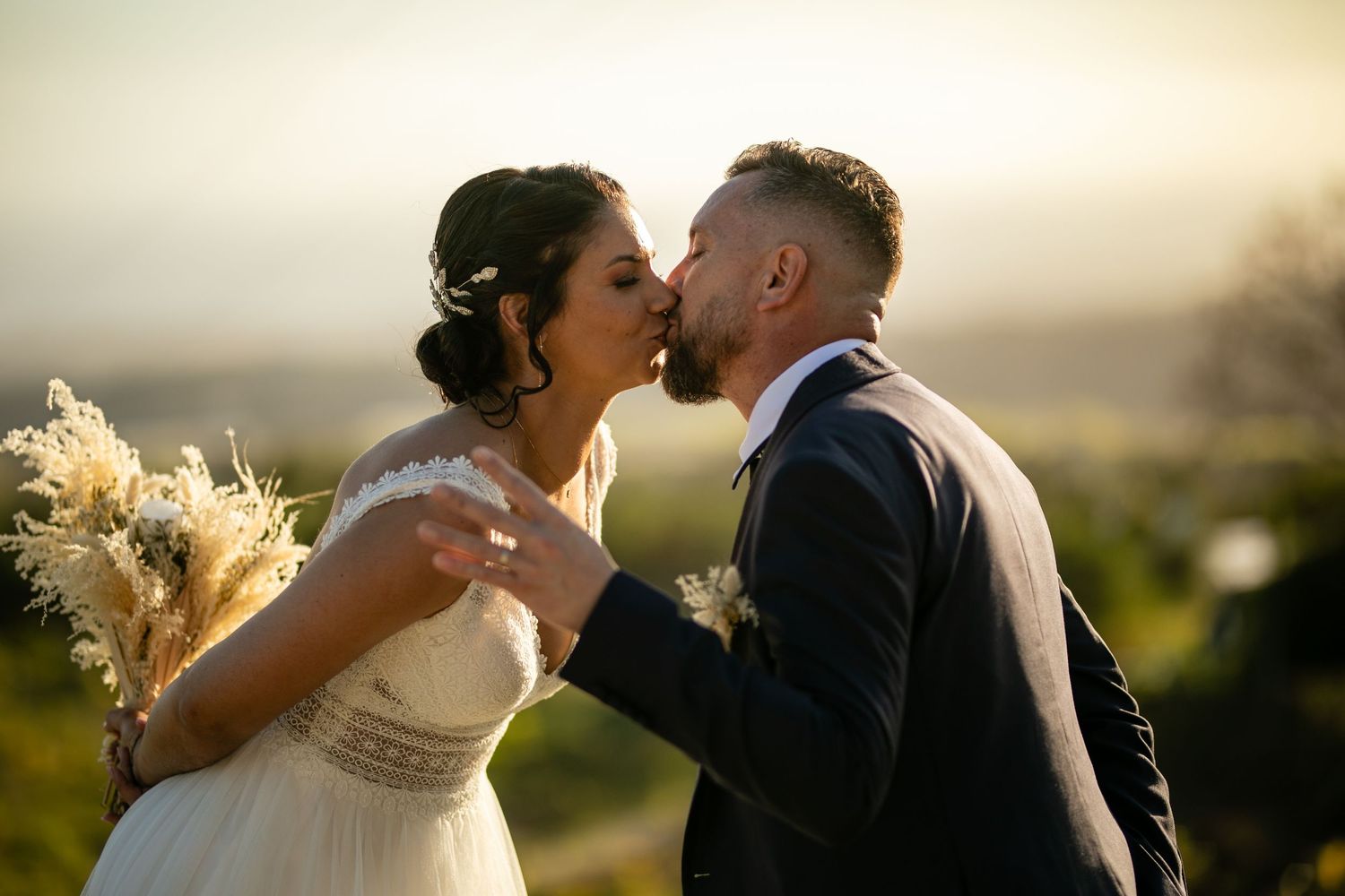 Romantic sunset wedding kiss between newlyweds in formal attire with pampas grass bouquet against golden sky.
