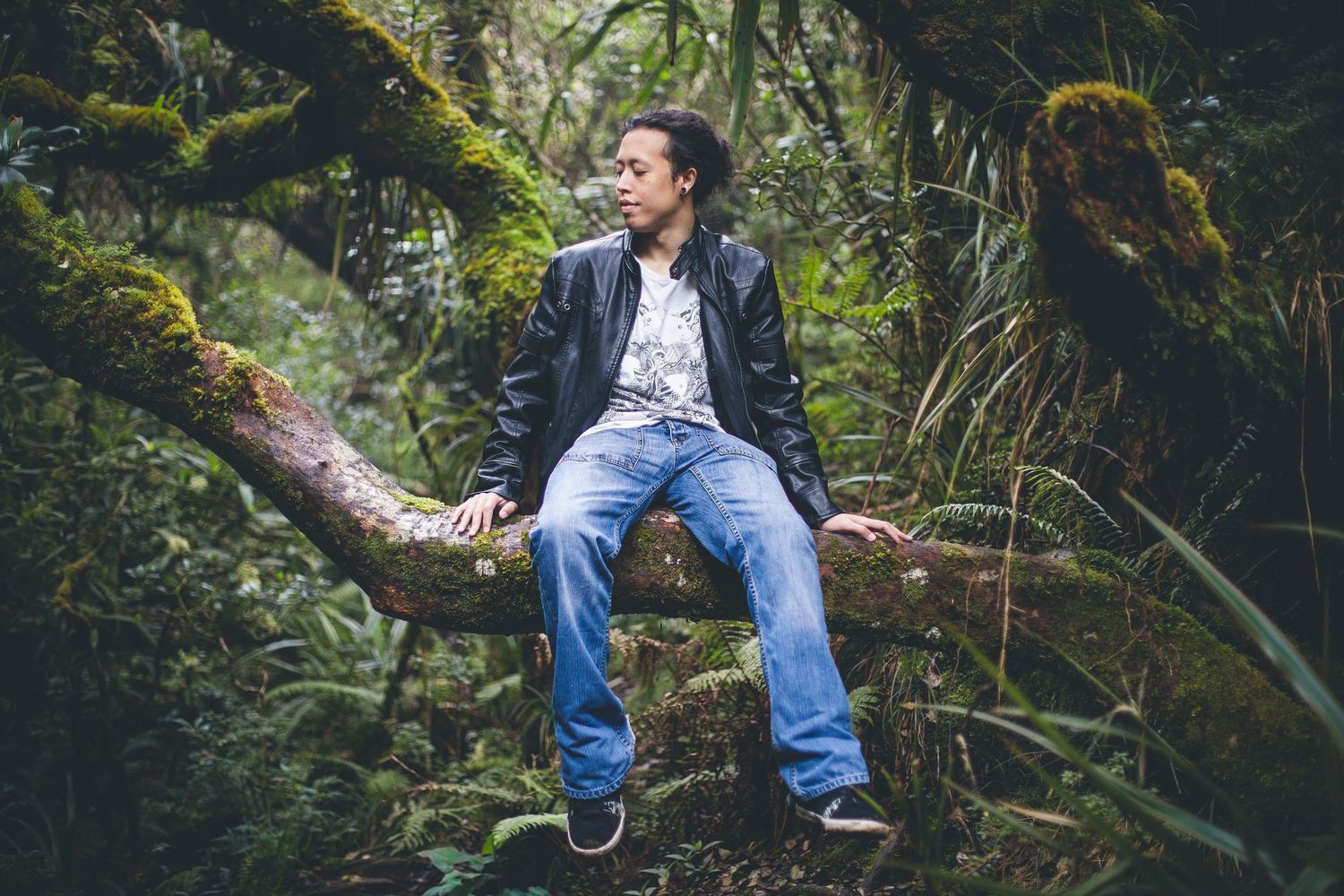 A casual portrait of someone in denim and leather jacket sitting on a mossy log in a lush forest.
