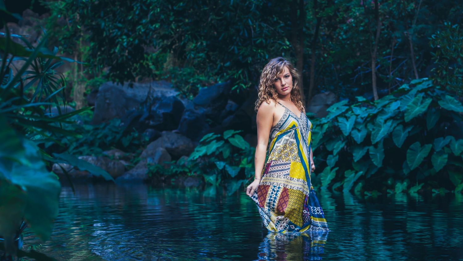 Person in summery dress standing in shallow water with lush forest background.