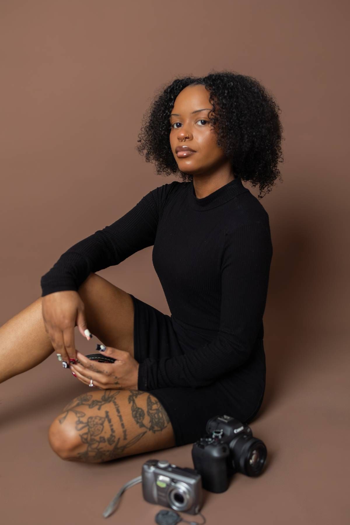 Young Black woman with curly hair in black dress sits on floor with tattoos and cameras nearby on brown background.