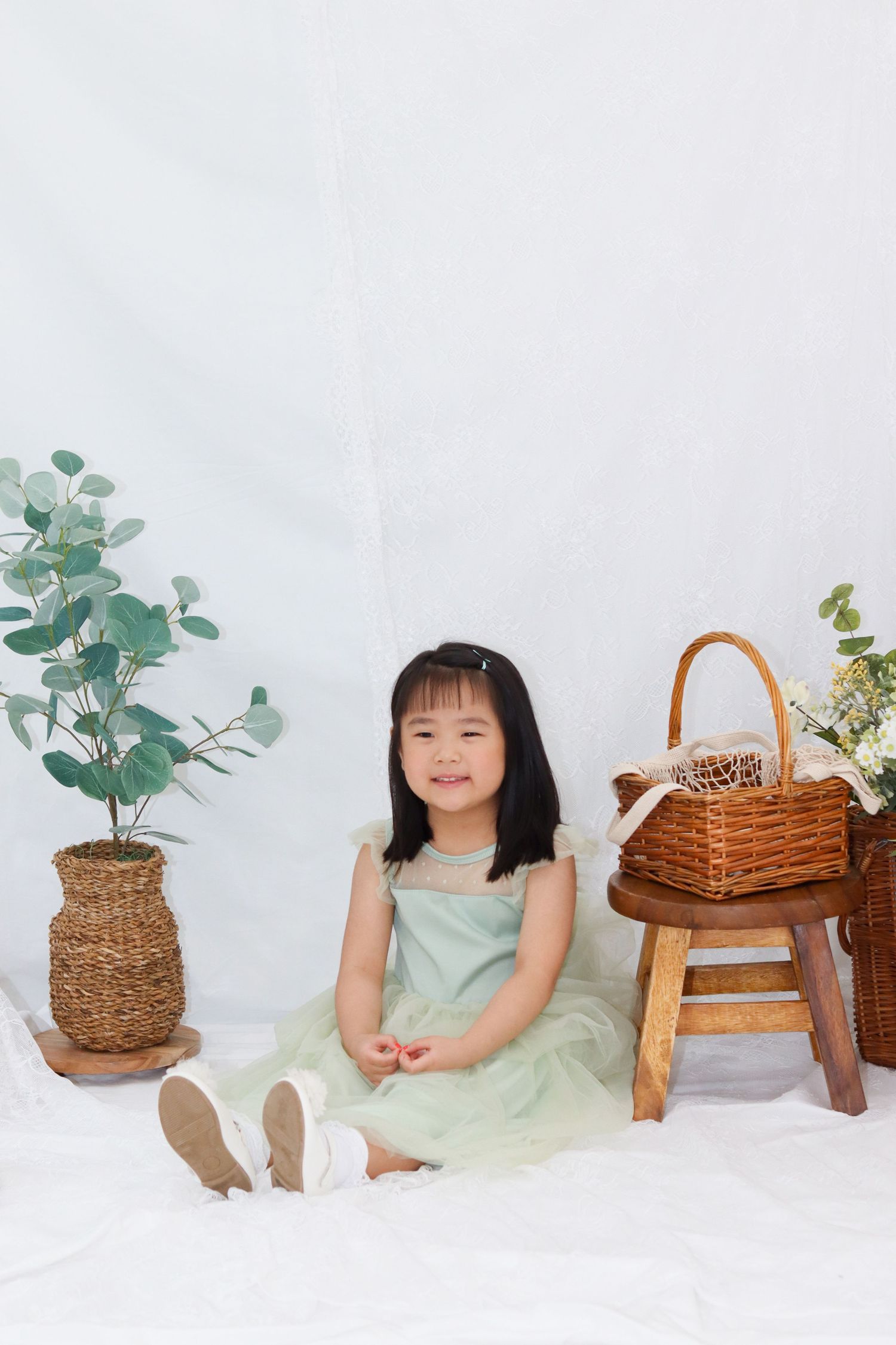 Studio portrait of child in mint green dress sitting on white floor with wicker basket and plants.