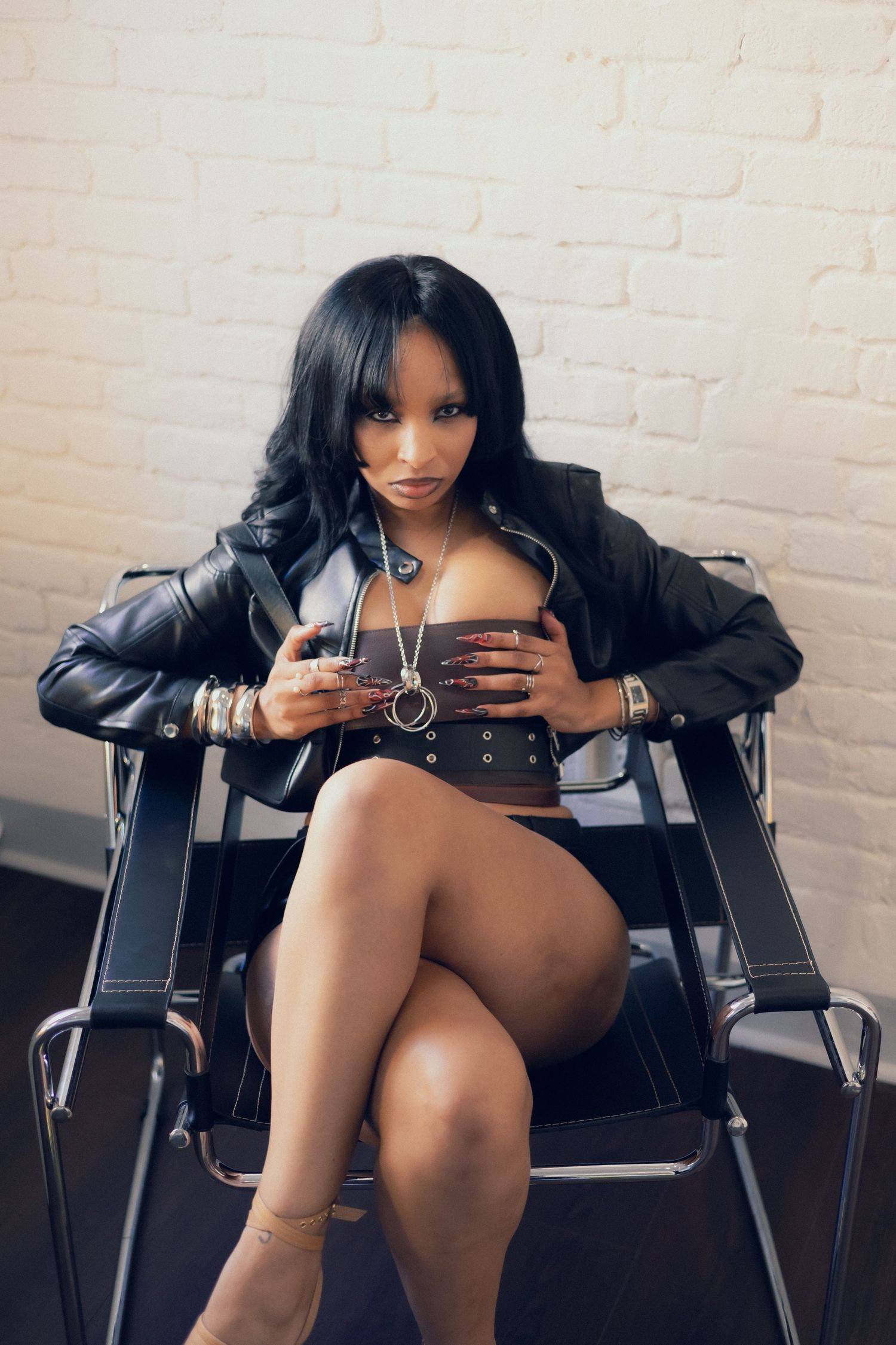 Woman in black leather jacket and mini skirt seated in chair, posing confidently against white brick wall.