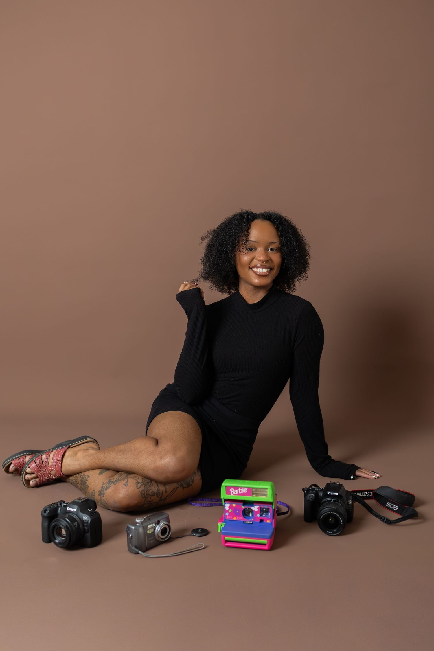 Smiling young woman in black dress sitting on brown studio floor surrounded by vintage and modern cameras.
