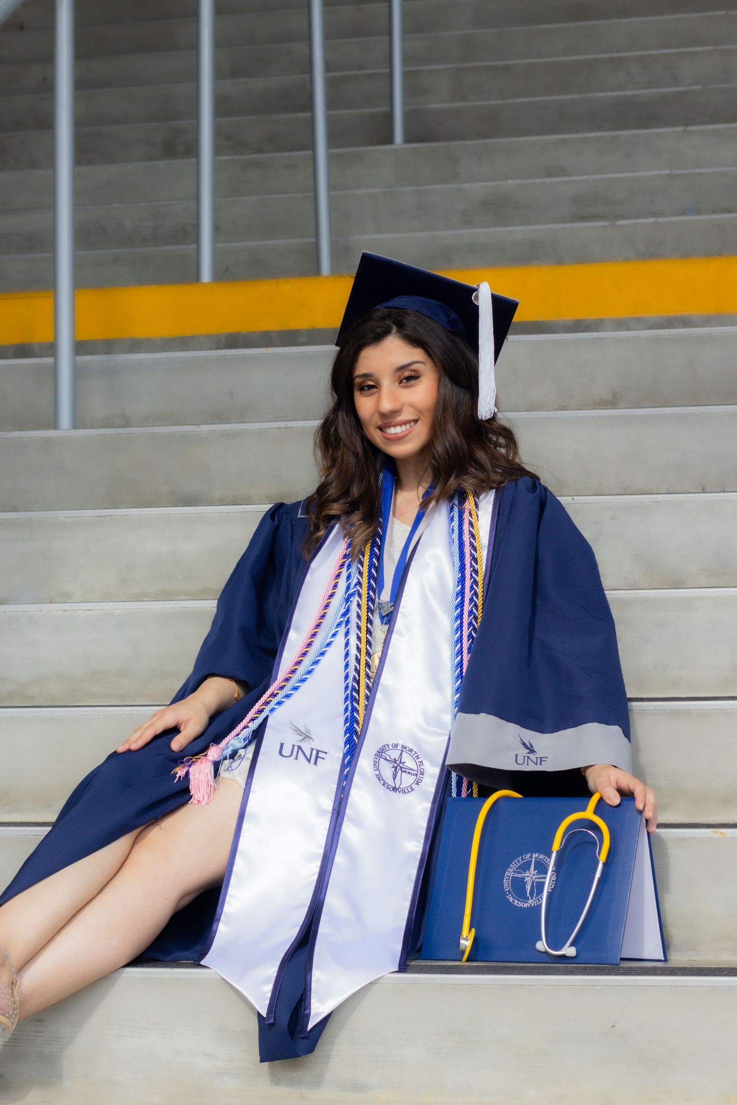 Graduate wearing blue cap and gown with red and white stole posing on concrete steps with stethoscope.