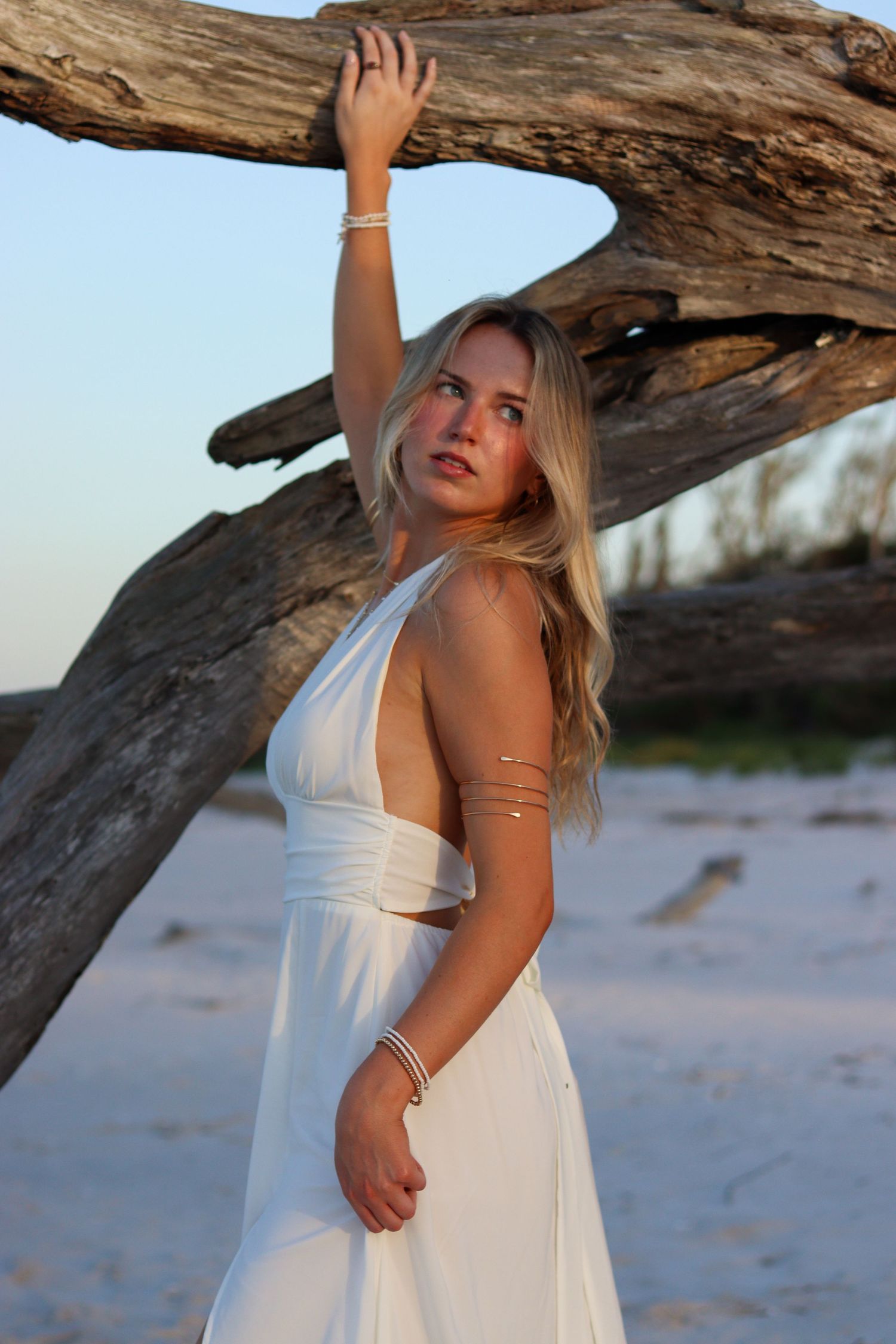 Beach photoshoot featuring model in white dress posing with large driftwood on sandy shore.