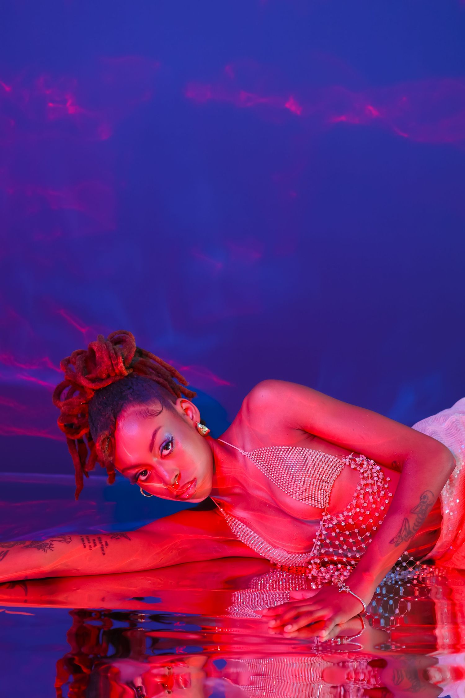 Artistic portrait with model lying down in sparkly dress under red and blue lighting.