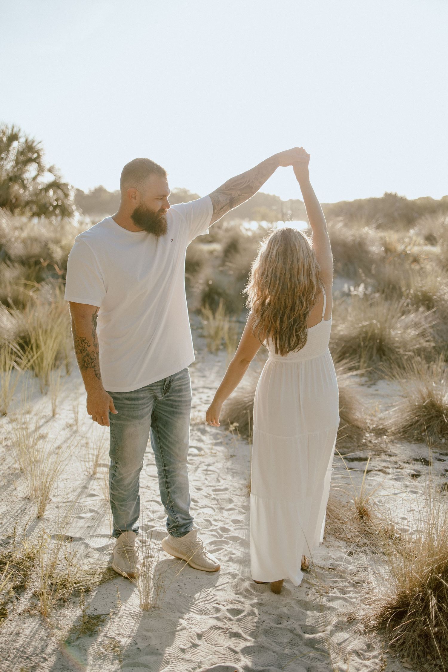 A couple dances together on a sandy beach during golden hour with coastal grasses in the background.