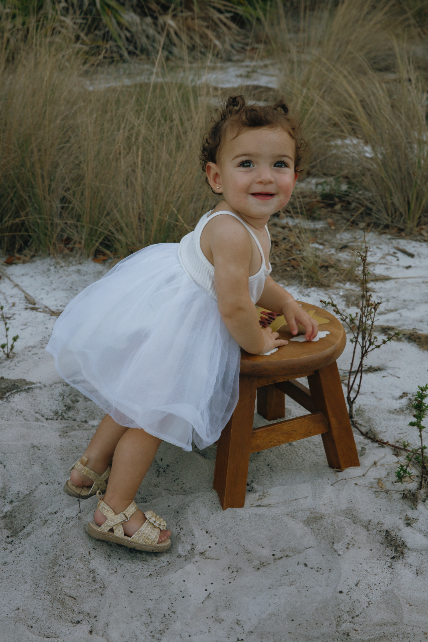 Child in white dress sits thoughtfully on wooden stool against sandy natural backdrop.