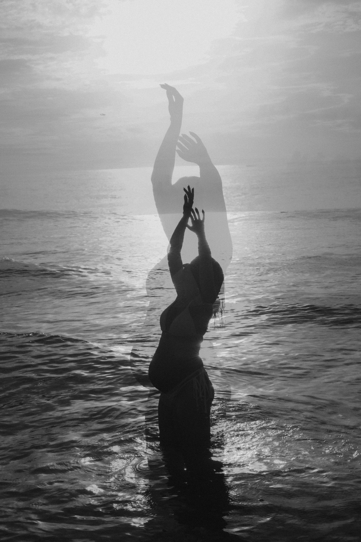 Silhouette of a figure reaching up with both arms while standing in ocean water at sunset creating dramatic contrast.