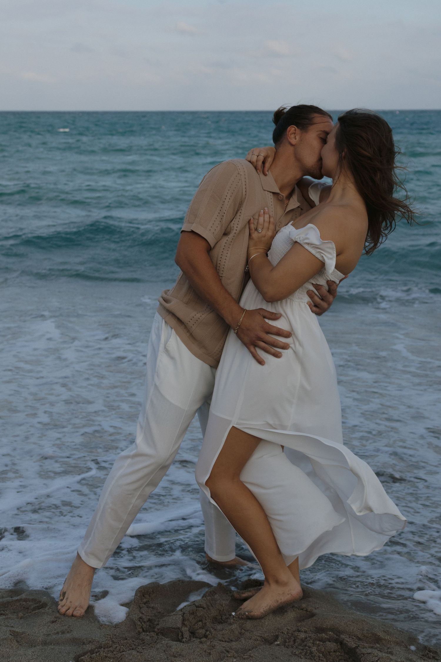 A couple in white clothing dances intimately on a beach with ocean waves in the background.