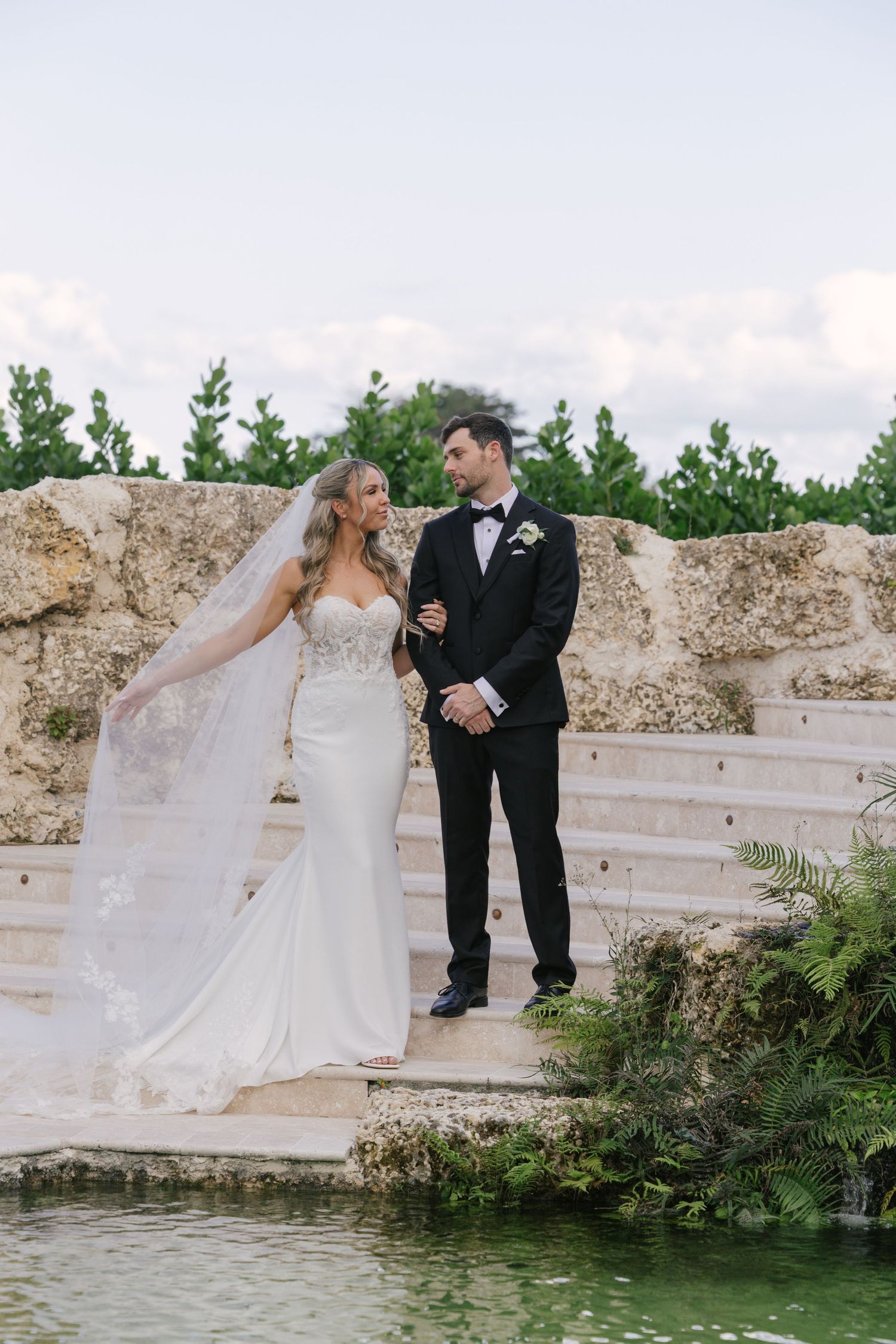 A newlywed couple stands on stone steps beside a pond, with the bride wearing a flowing white gown and long veil.