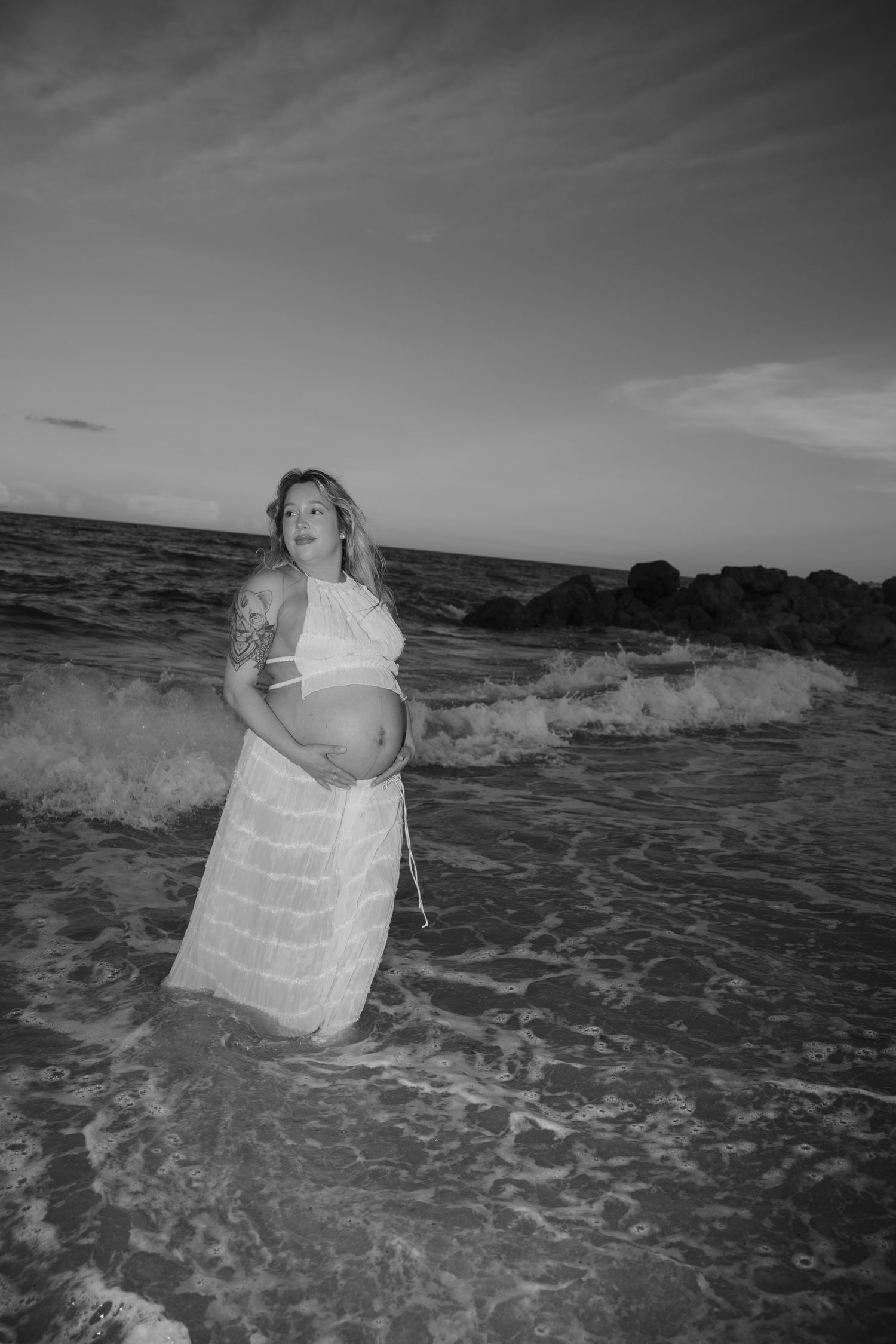 Black and white photo of pregnant woman standing in ocean surf wearing flowing white dress.