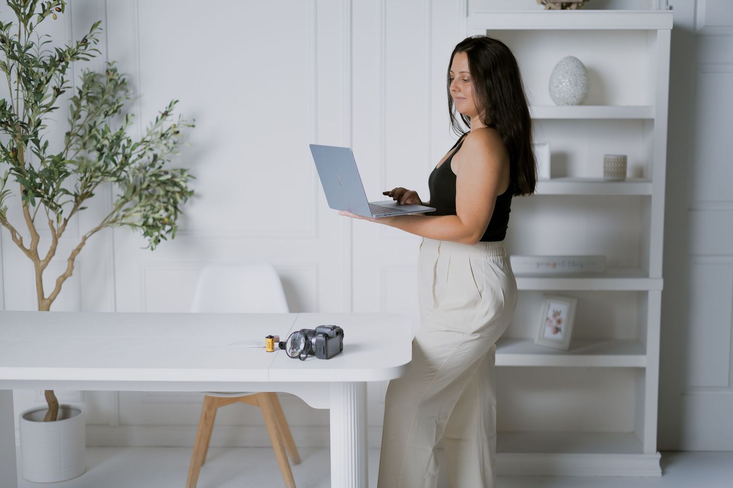 Woman in black top and beige pants stands against white wall using laptop in modern minimalist home office.