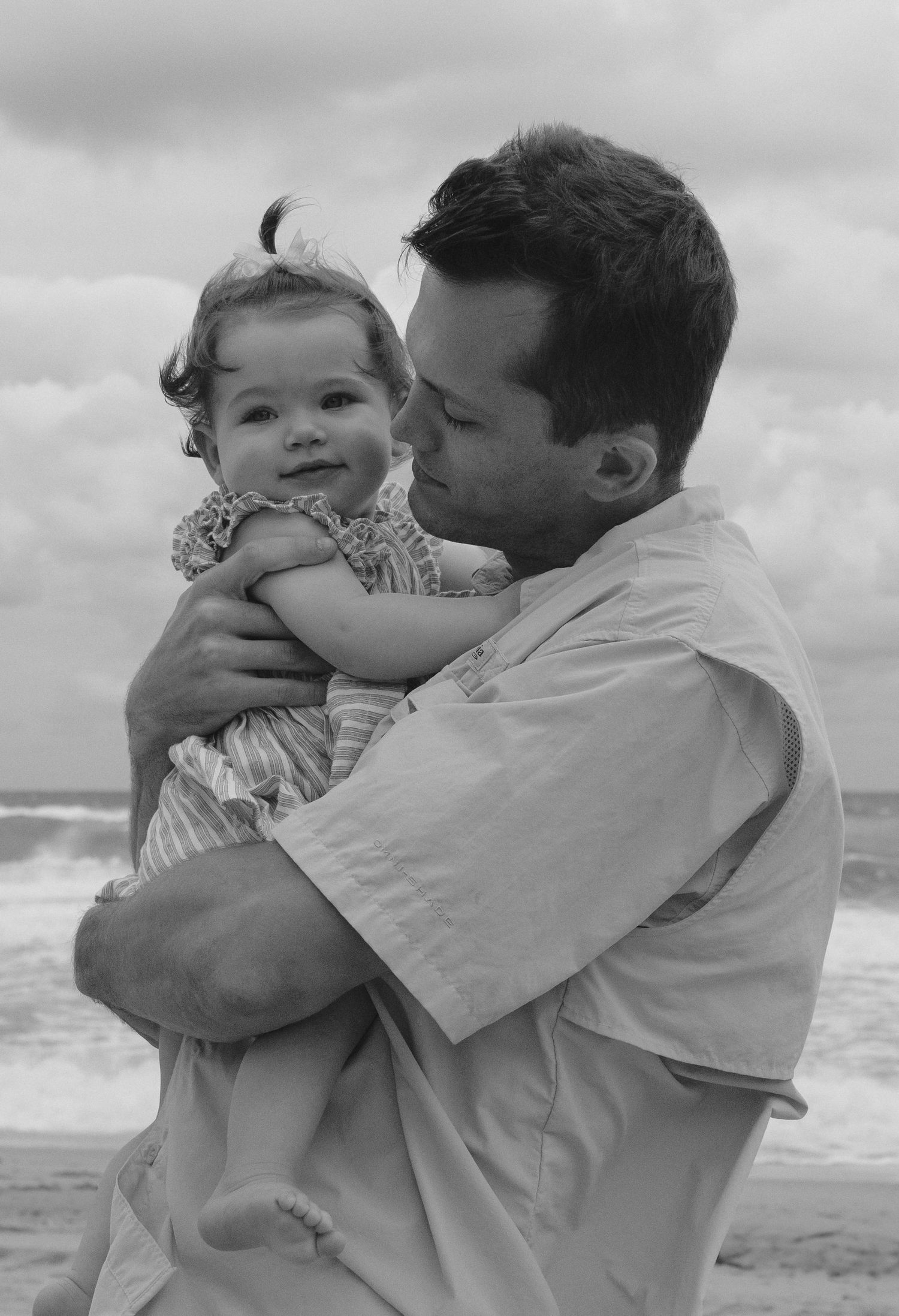 A tender black and white beach portrait of a parent holding a smiling toddler in a floral dress against an ocean backdrop.