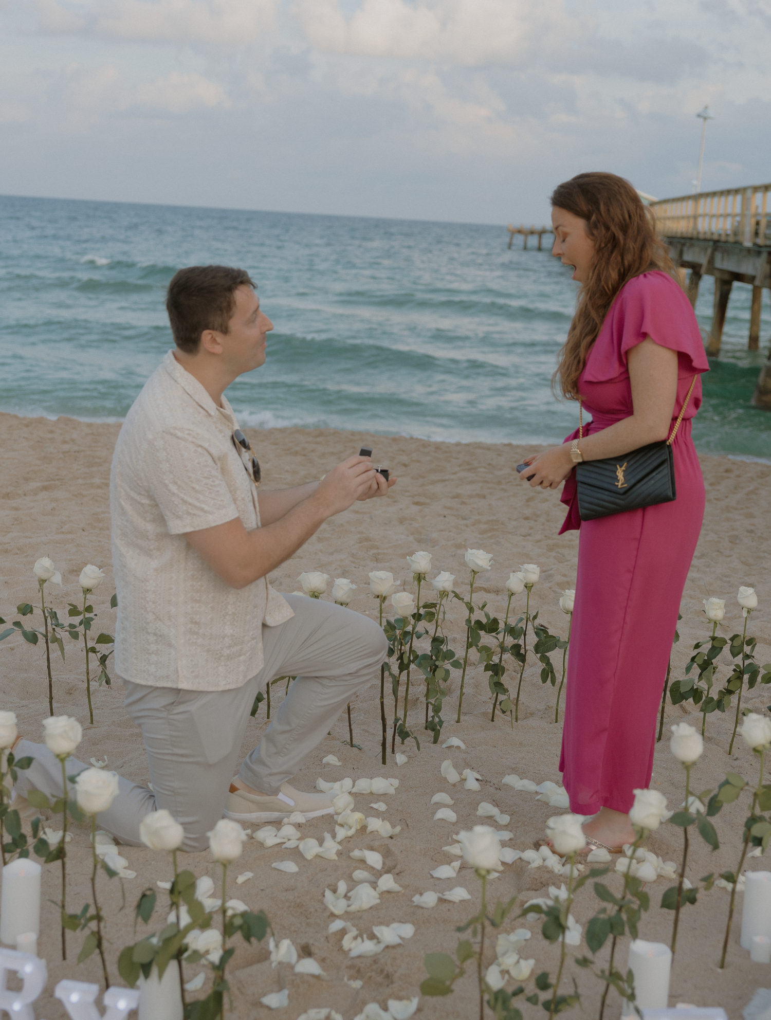 A romantic beach proposal scene with white rose petals and candles arranged on the sand at sunset near a pier.