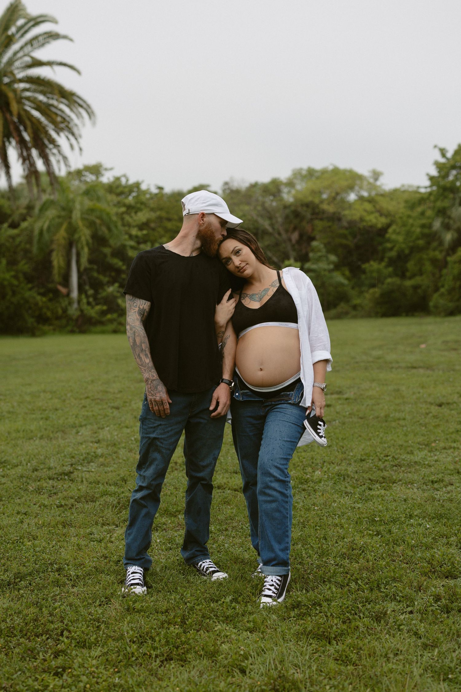 Two people in casual clothing standing close together on a grassy field with palm trees in the background.