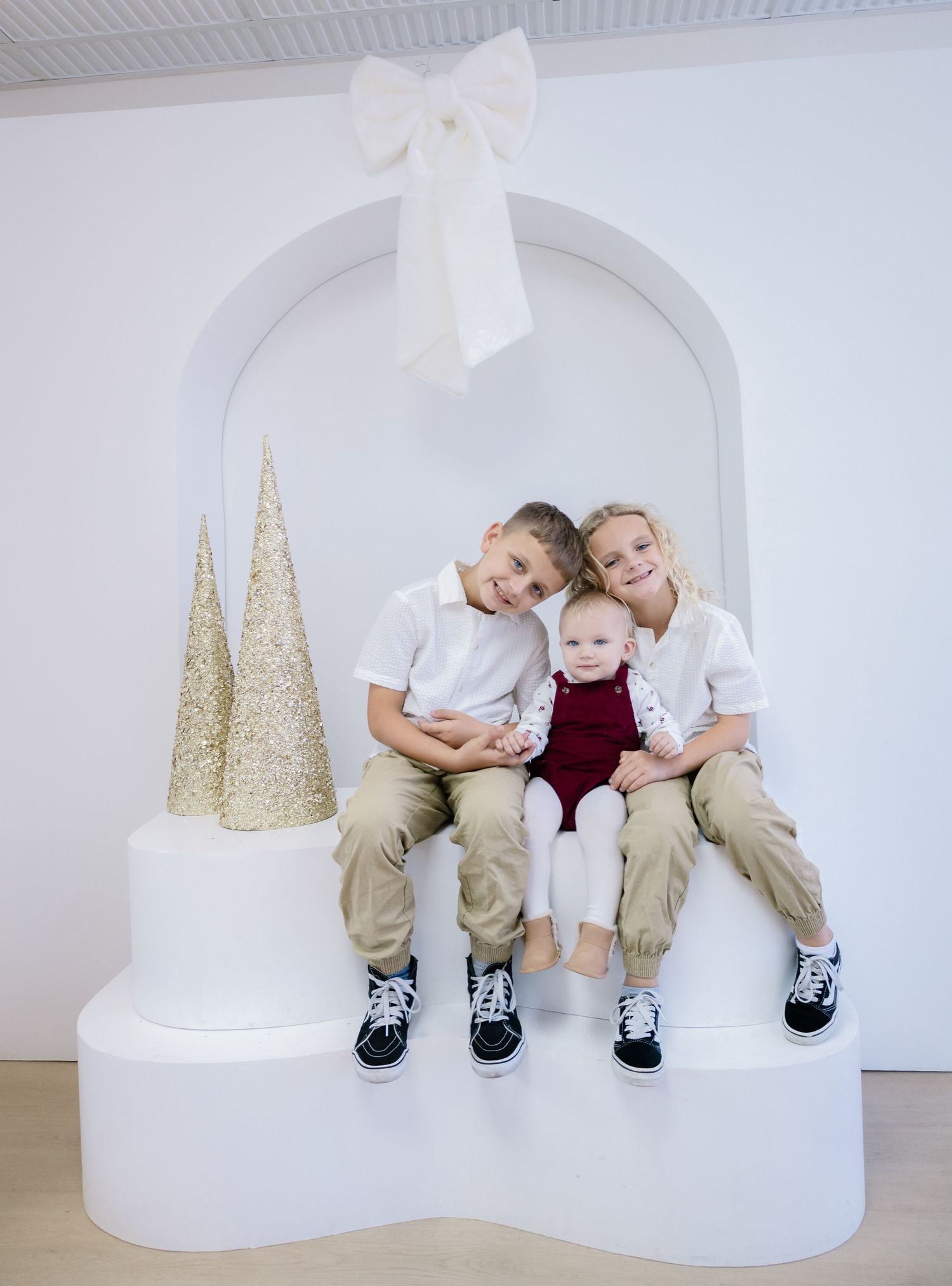 Children sitting together on a white curved platform with gold glitter Christmas trees in the background for holiday portraits.
