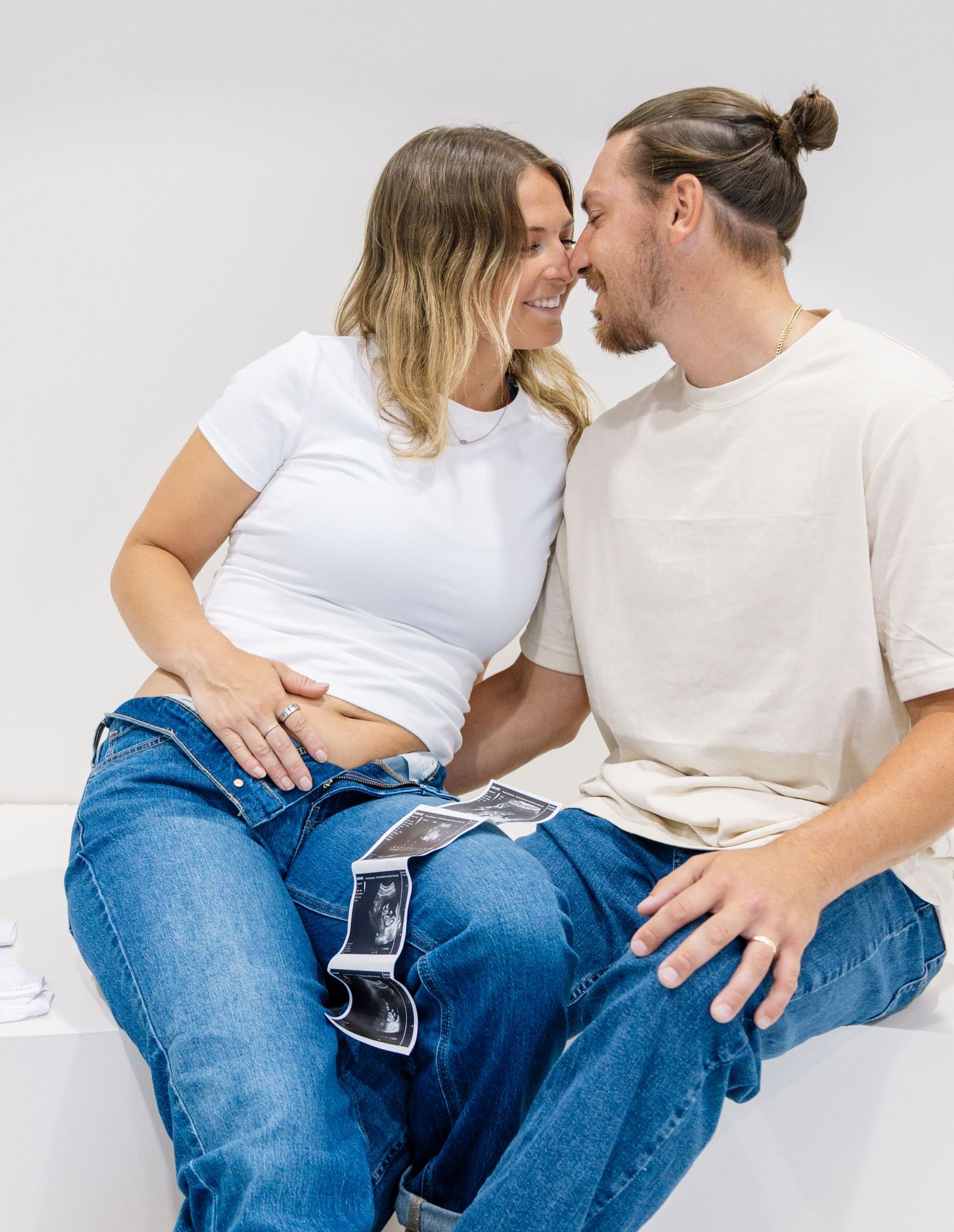A pregnant couple in white tops and blue jeans share a tender moment with ultrasound photos on their laps.