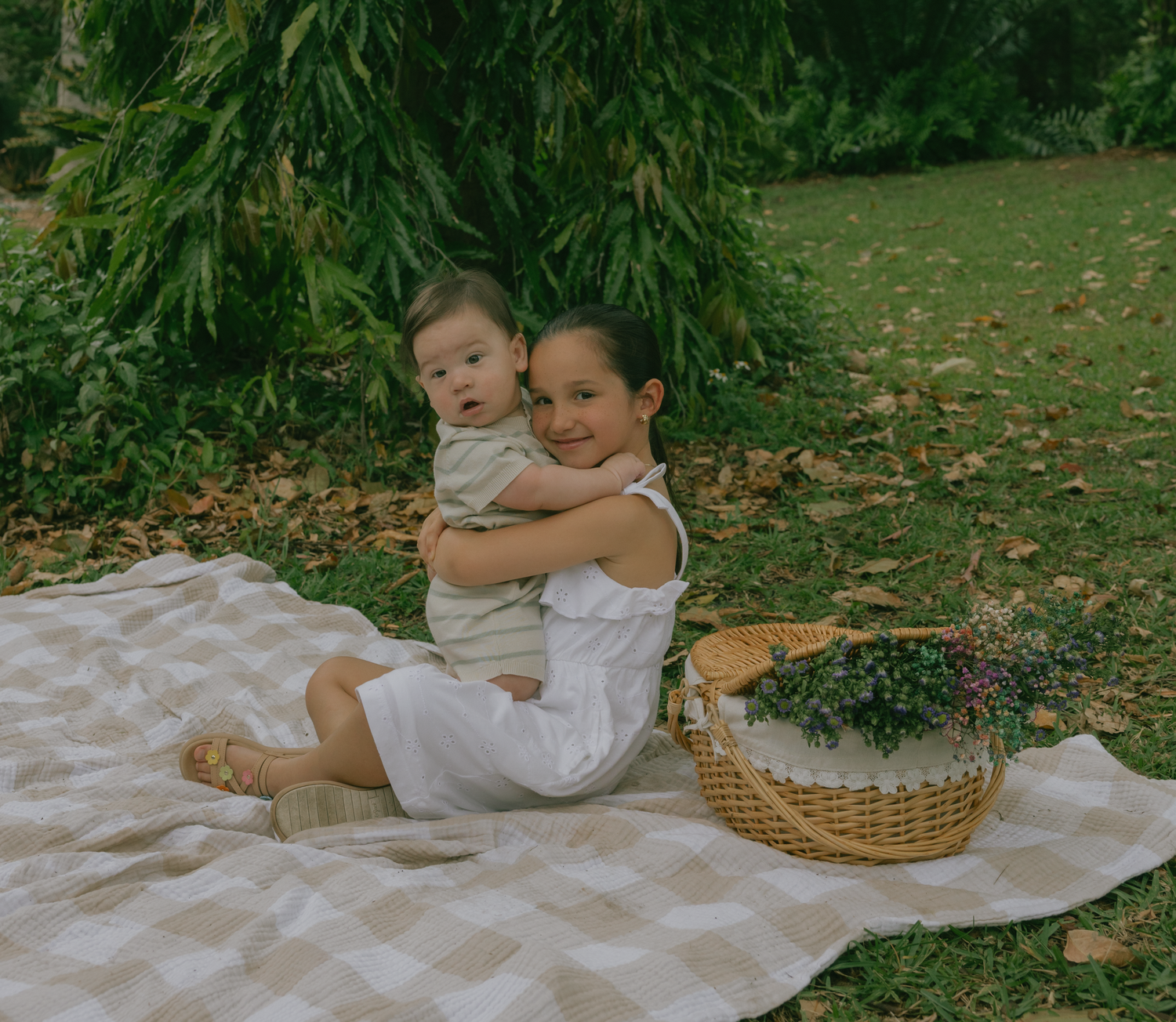 Two children hugging while sitting on checkered blanket next to picnic basket.