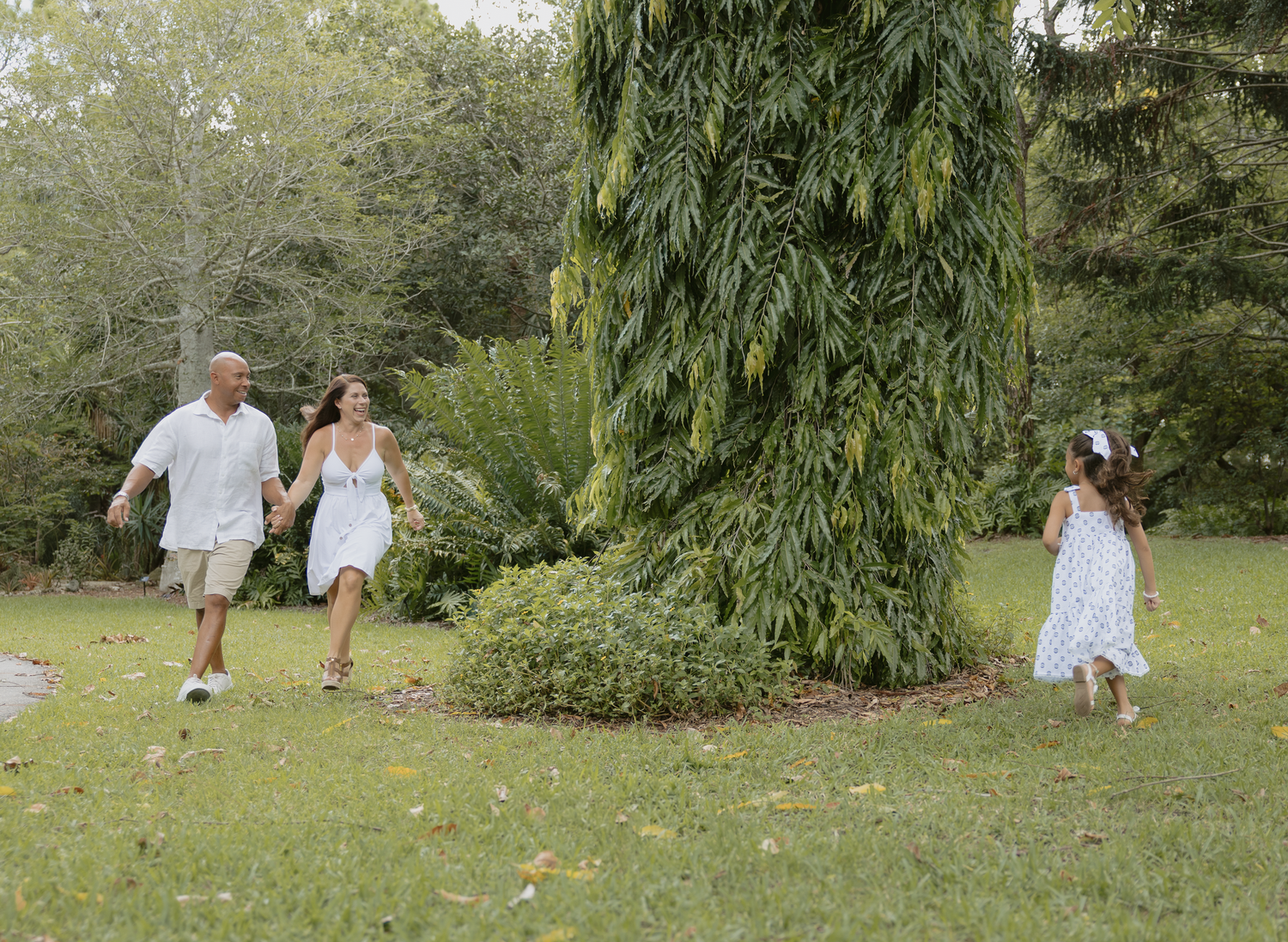 People in white clothing walk and play together on a grassy lawn surrounded by lush greenery.