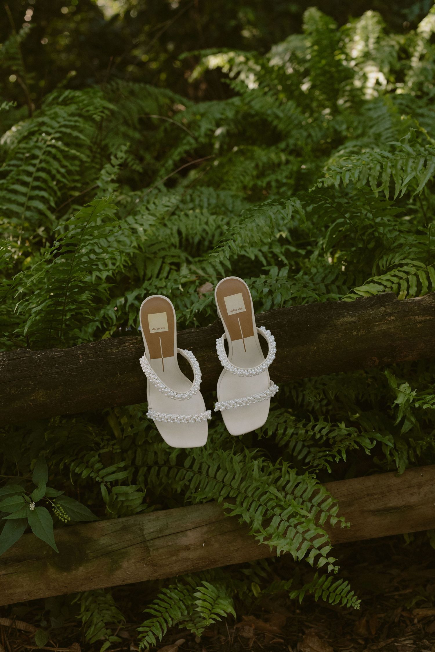 White wedding sandals with delicate straps rest on a wooden fence surrounded by lush green ferns in a garden setting.