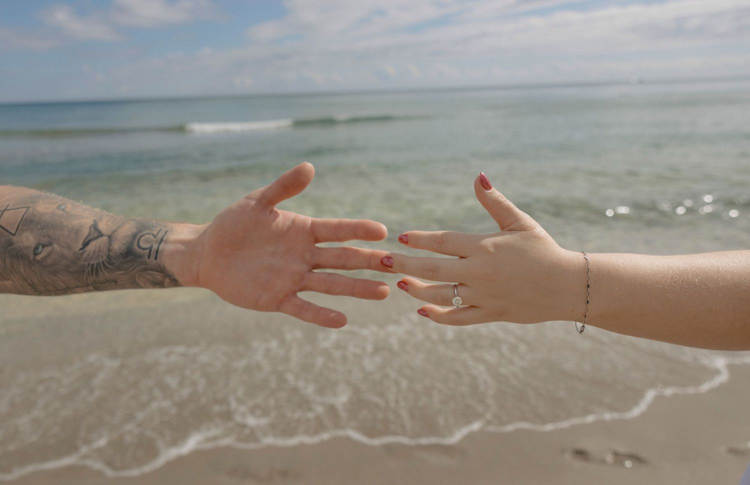 A hand reaches to touch the waves gently rolling onto a pristine sandy beach on a cloudy day.