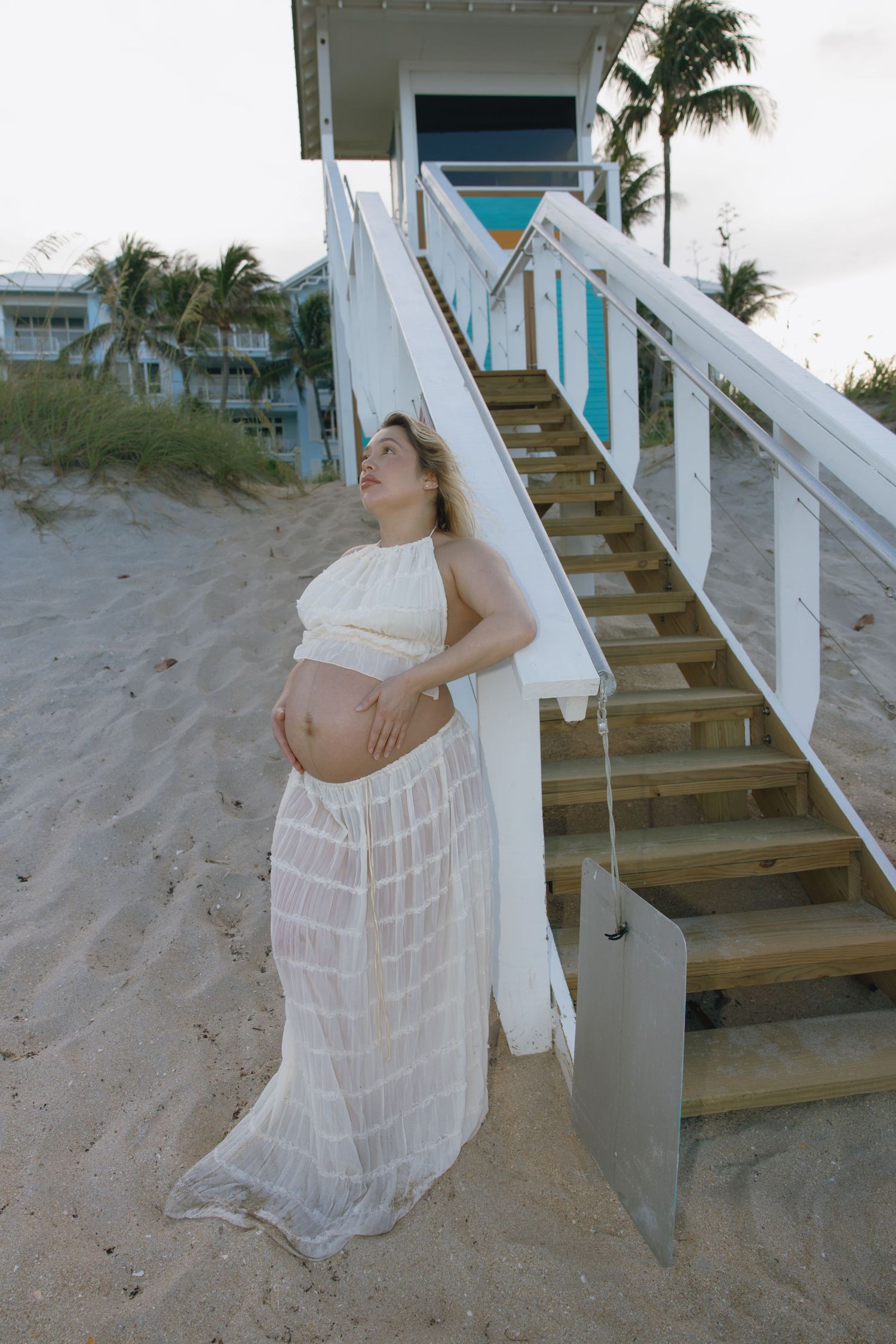 Woman in white sheer dress poses dramatically on sandy beach stairs near palm trees and turquoise building.