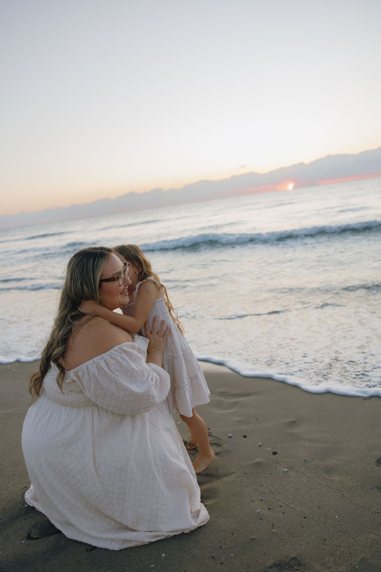 An intimate moment between two people in white dresses on a beach as waves roll in at sunset.