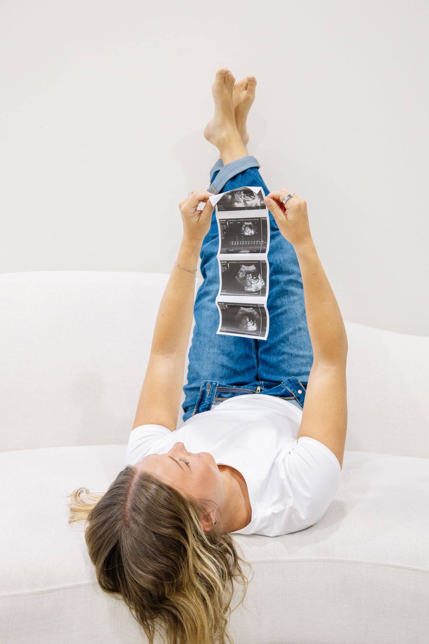 Pregnant woman lying on sofa holding ultrasound scan images up while wearing white shirt and blue jeans.