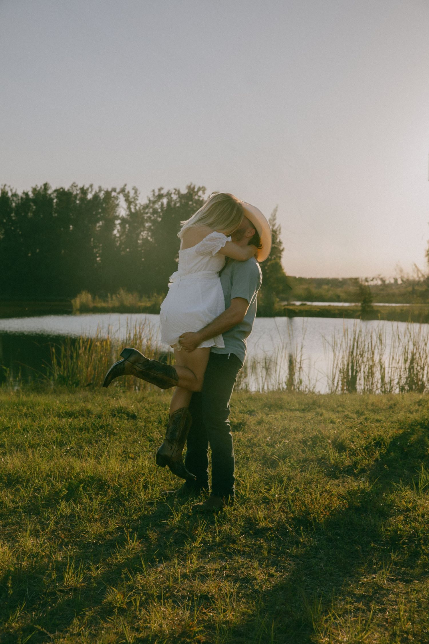 A couple dances and embraces in a golden field at sunset near a serene lake.