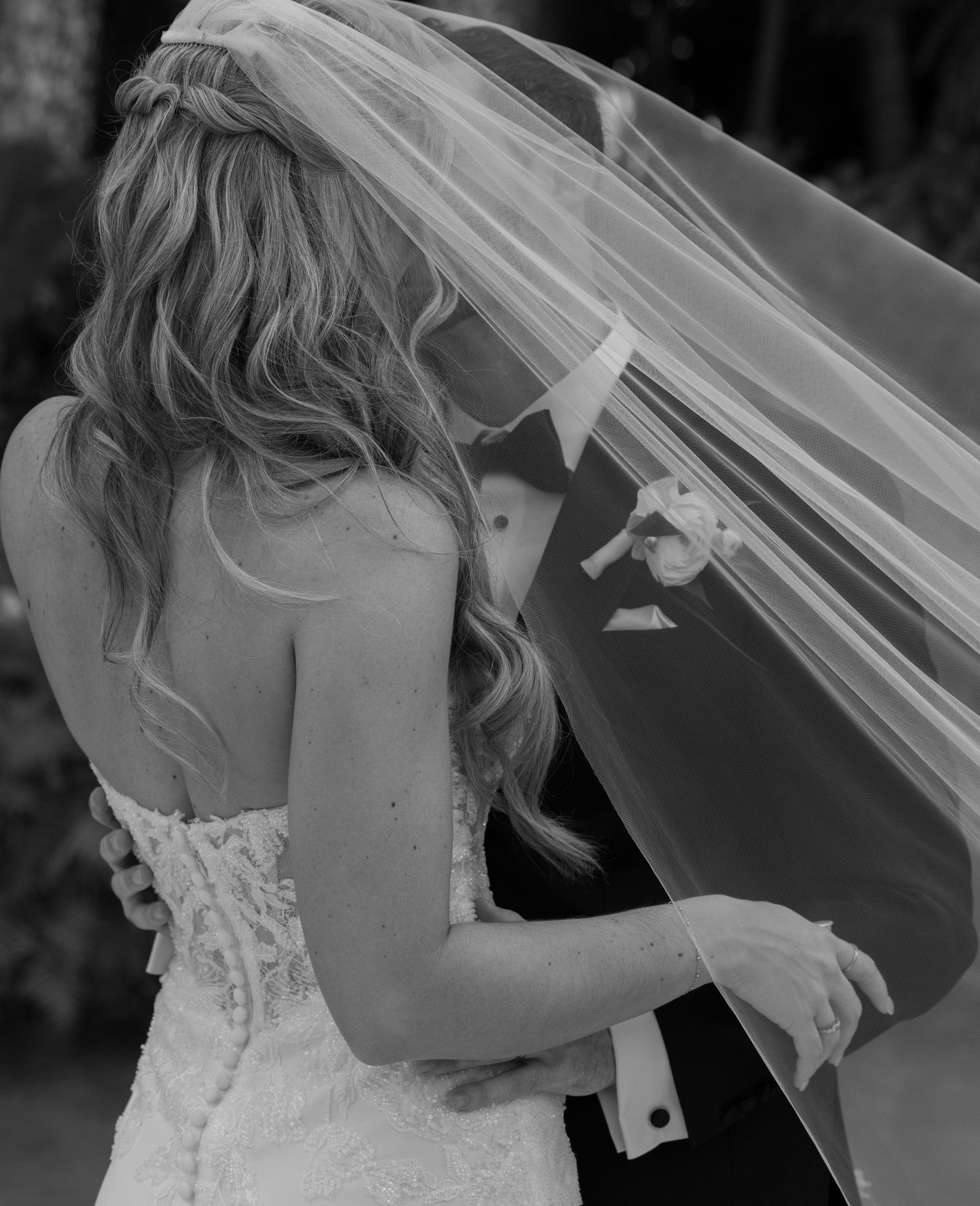 Black and white wedding photo showing a bride in a lace dress with flowing veil embracing someone during a tender moment.