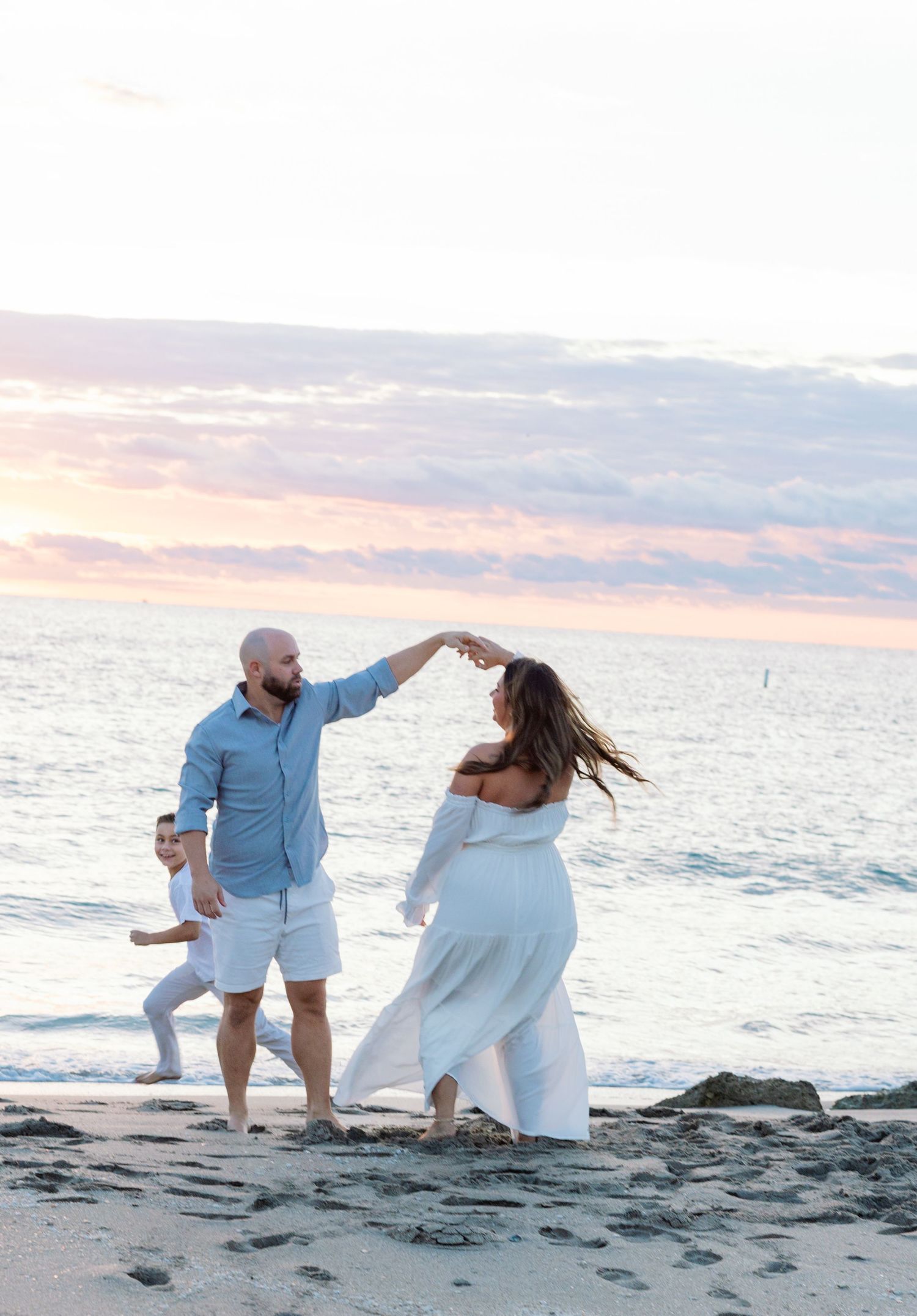 Couple dancing on beach at sunset, woman in white maternity dress, man in blue shirt, child running in background.