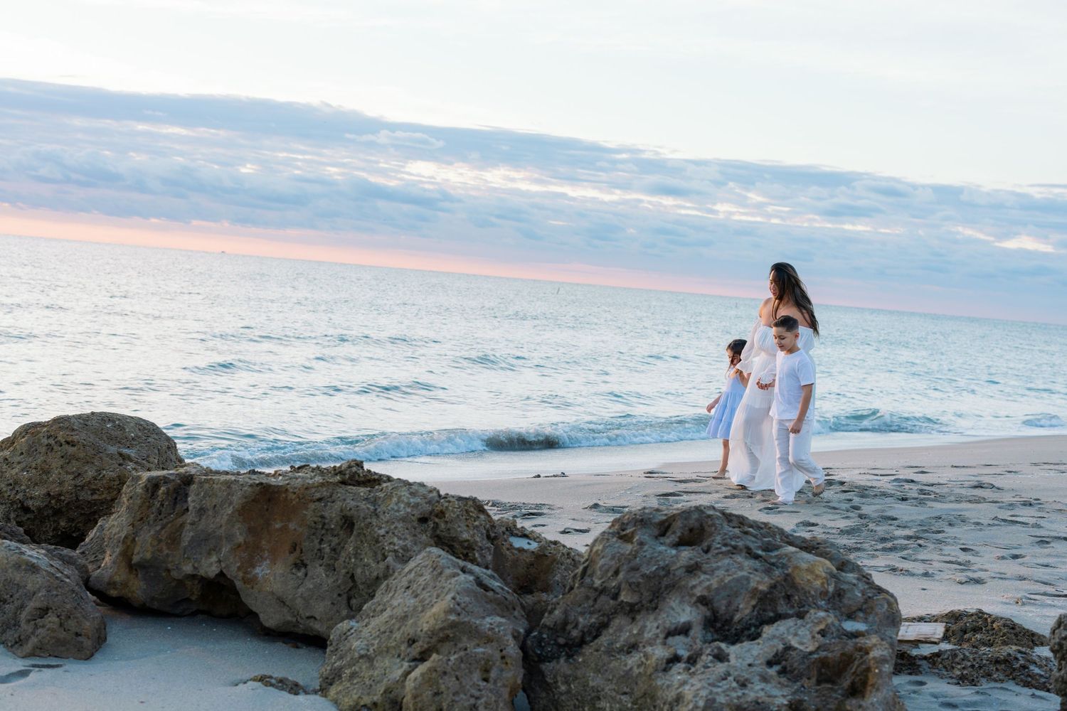 A mother and two children in white clothing stand on a sandy beach near rocks at sunset.