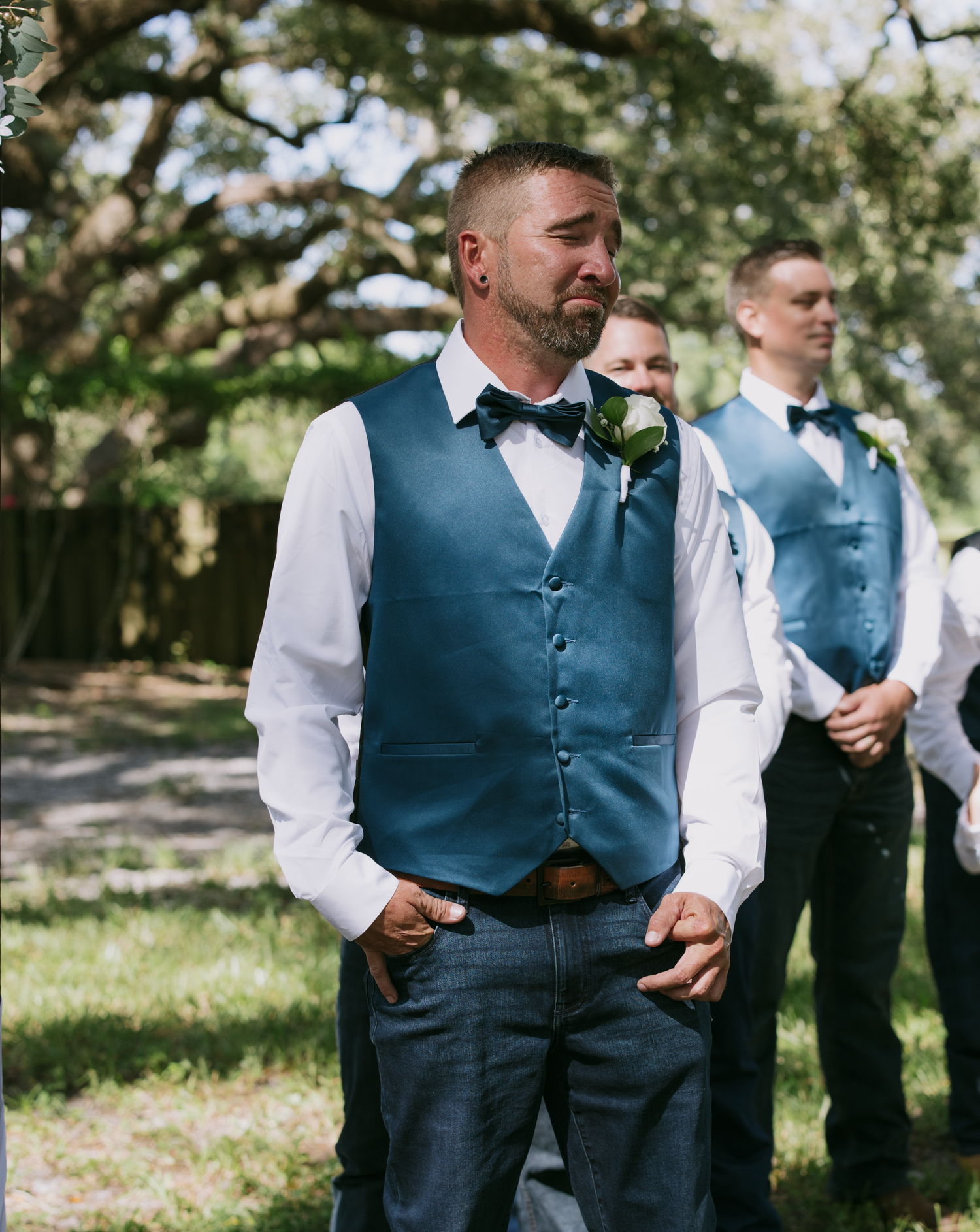 Groomsmen in navy blue vests and bow ties stand outdoors under trees during a formal wedding celebration.