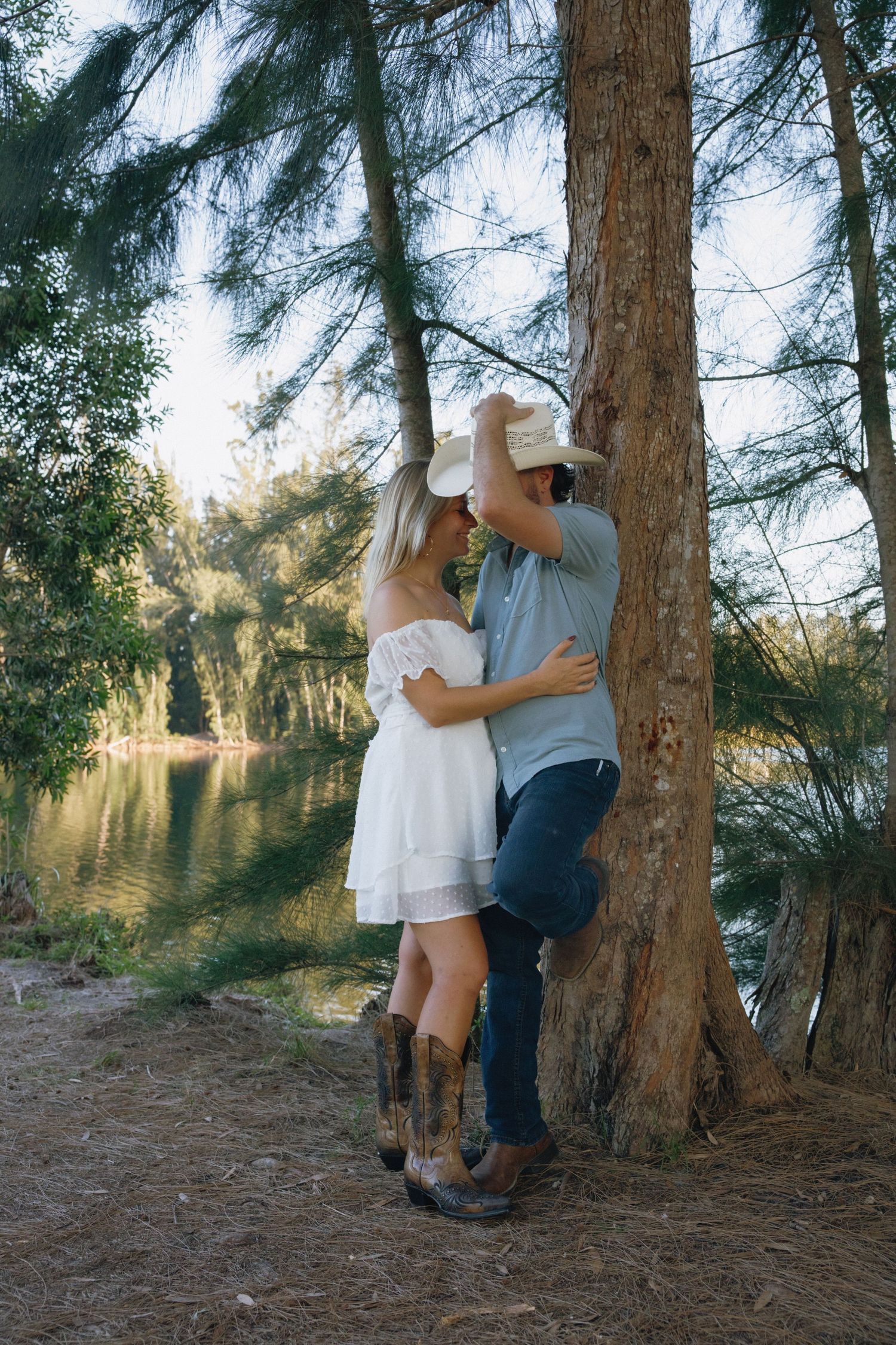 A romantic couple in cowboy boots embraces by a tree near a peaceful lake during golden hour.