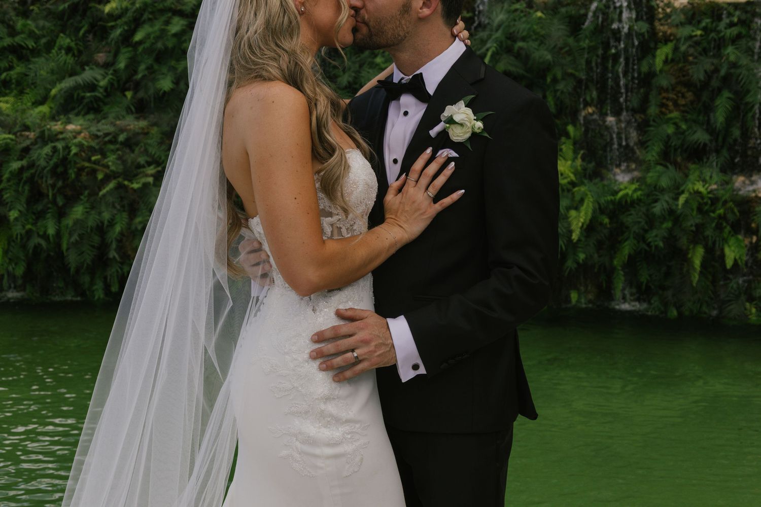 A couple shares a romantic kiss during their outdoor wedding ceremony by a lake surrounded by lush greenery.