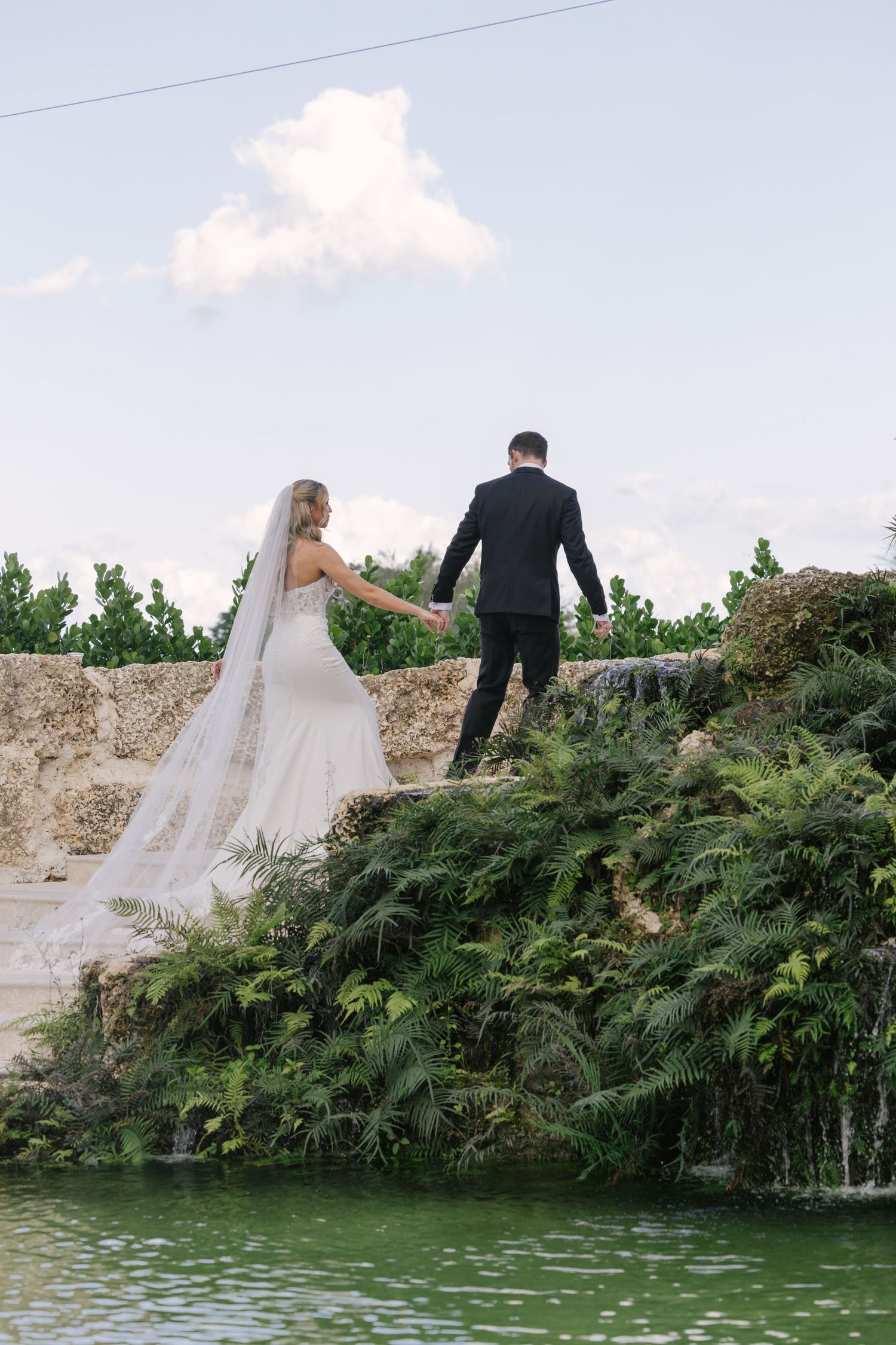 A couple in wedding attire walks along a scenic stone wall overlooking a tranquil pond surrounded by lush greenery.