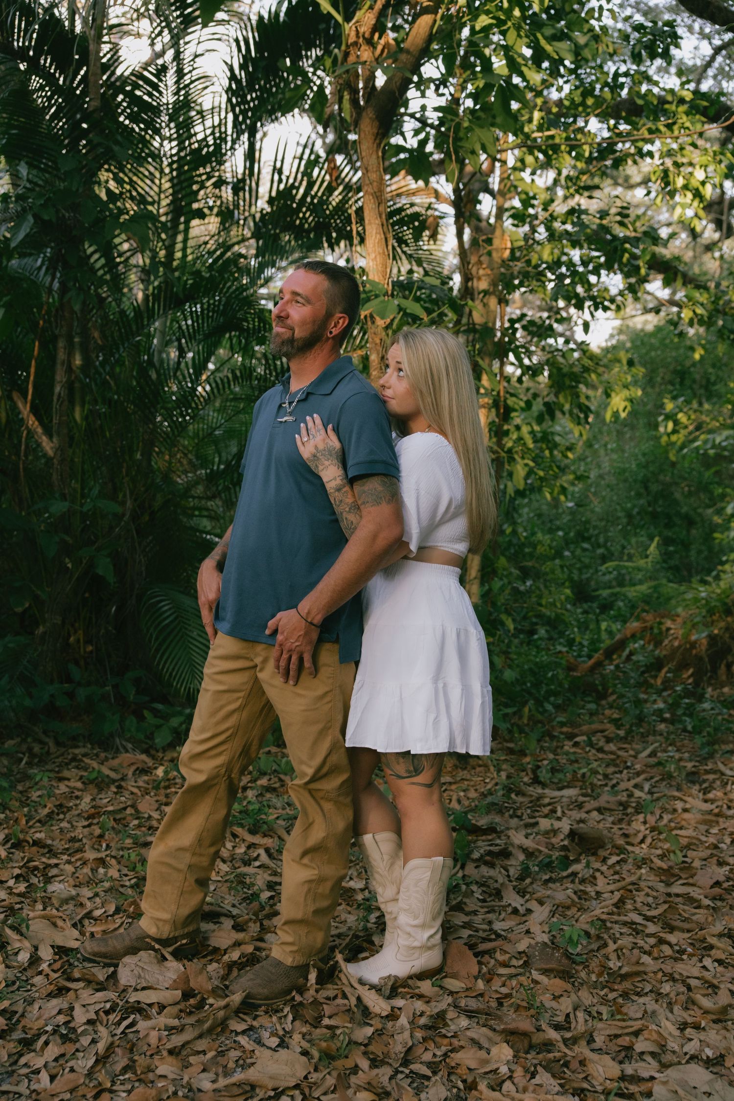 Romantic couple in casual clothing embrace while standing on fallen leaves in a wooded outdoor setting at sunset.