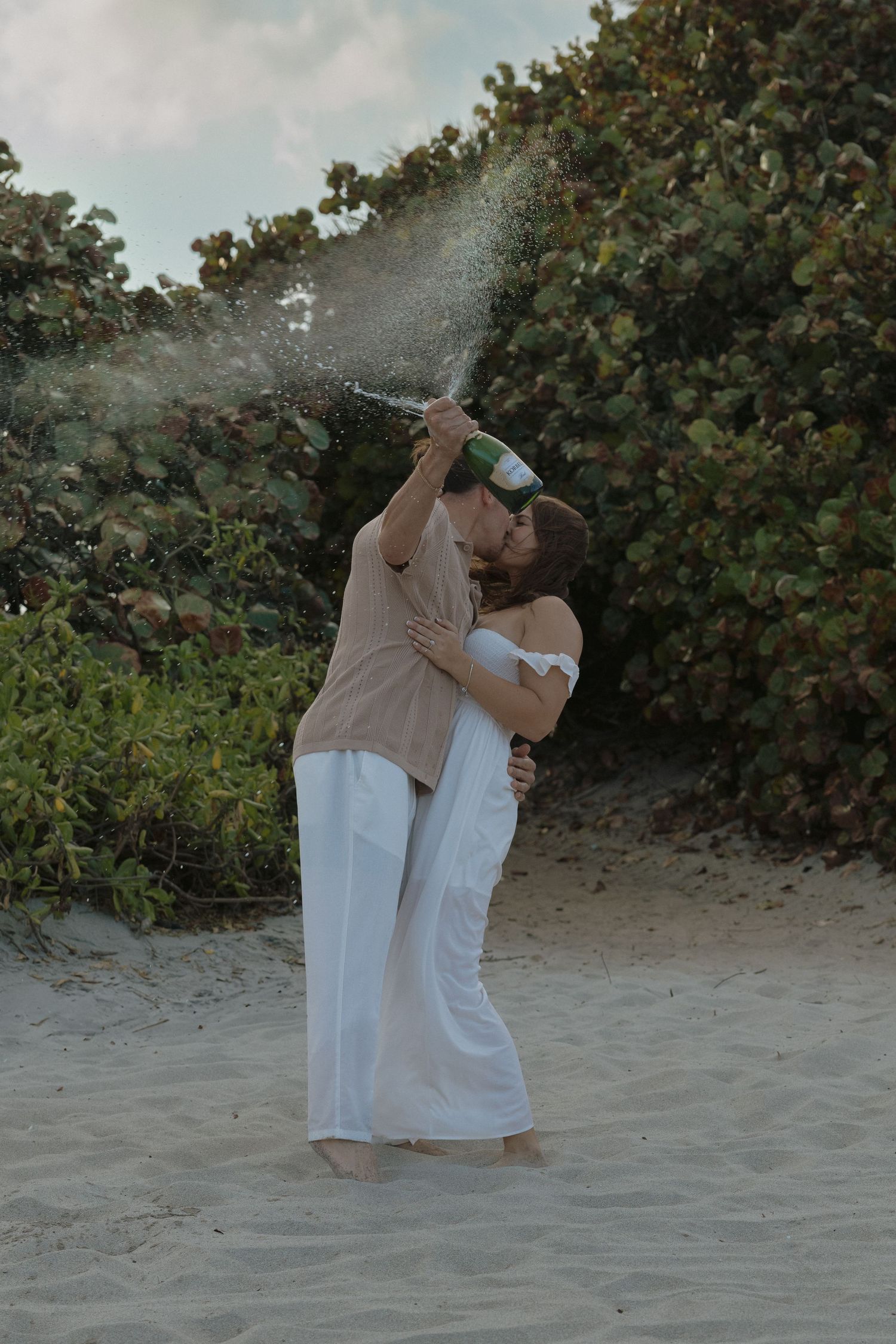A couple stands together on a sandy beach wearing white outfits, creating a romantic atmosphere at dusk.
