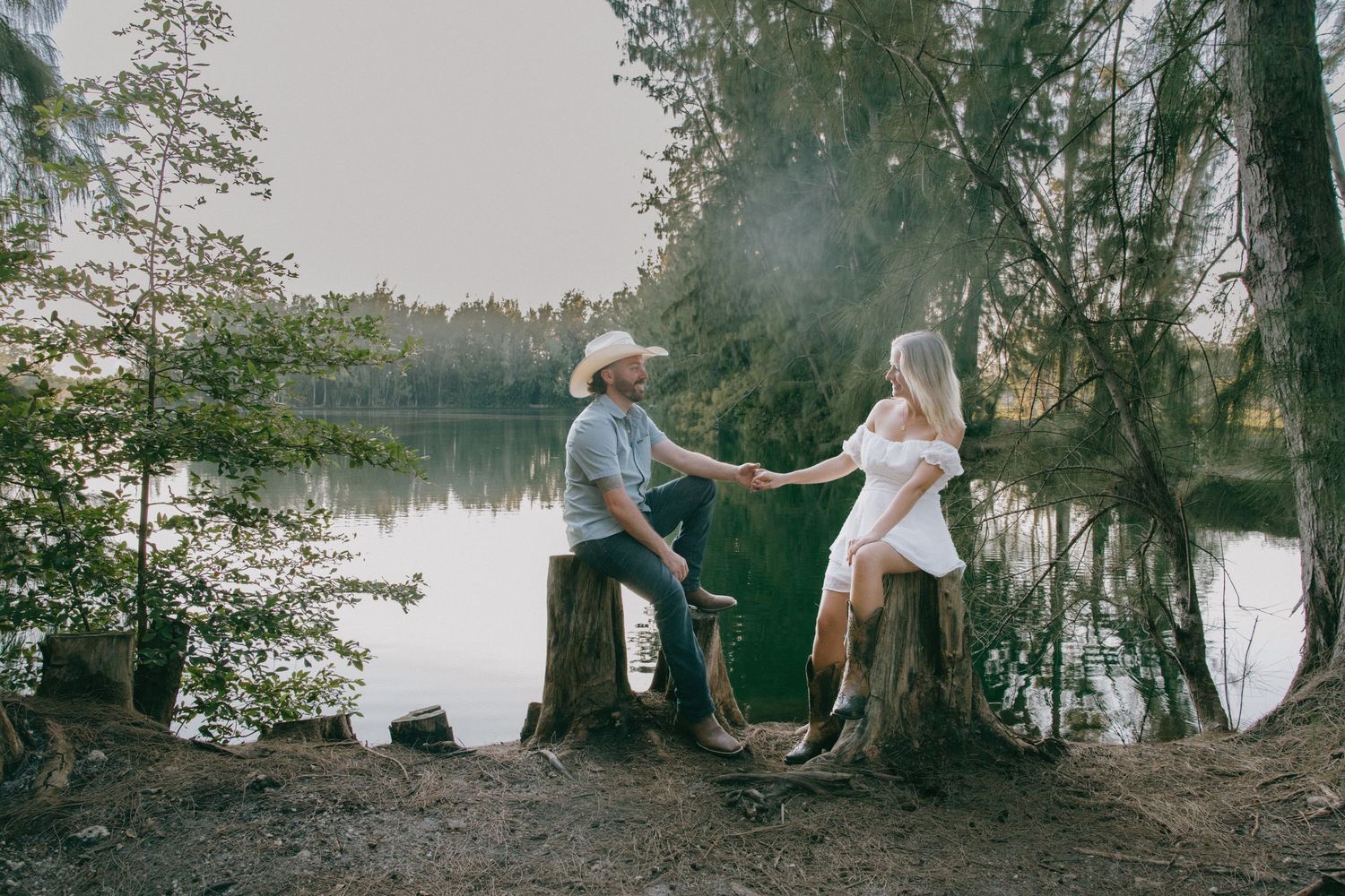 A couple enjoys a romantic moment by a serene lake with mist rising through trees at sunset.
