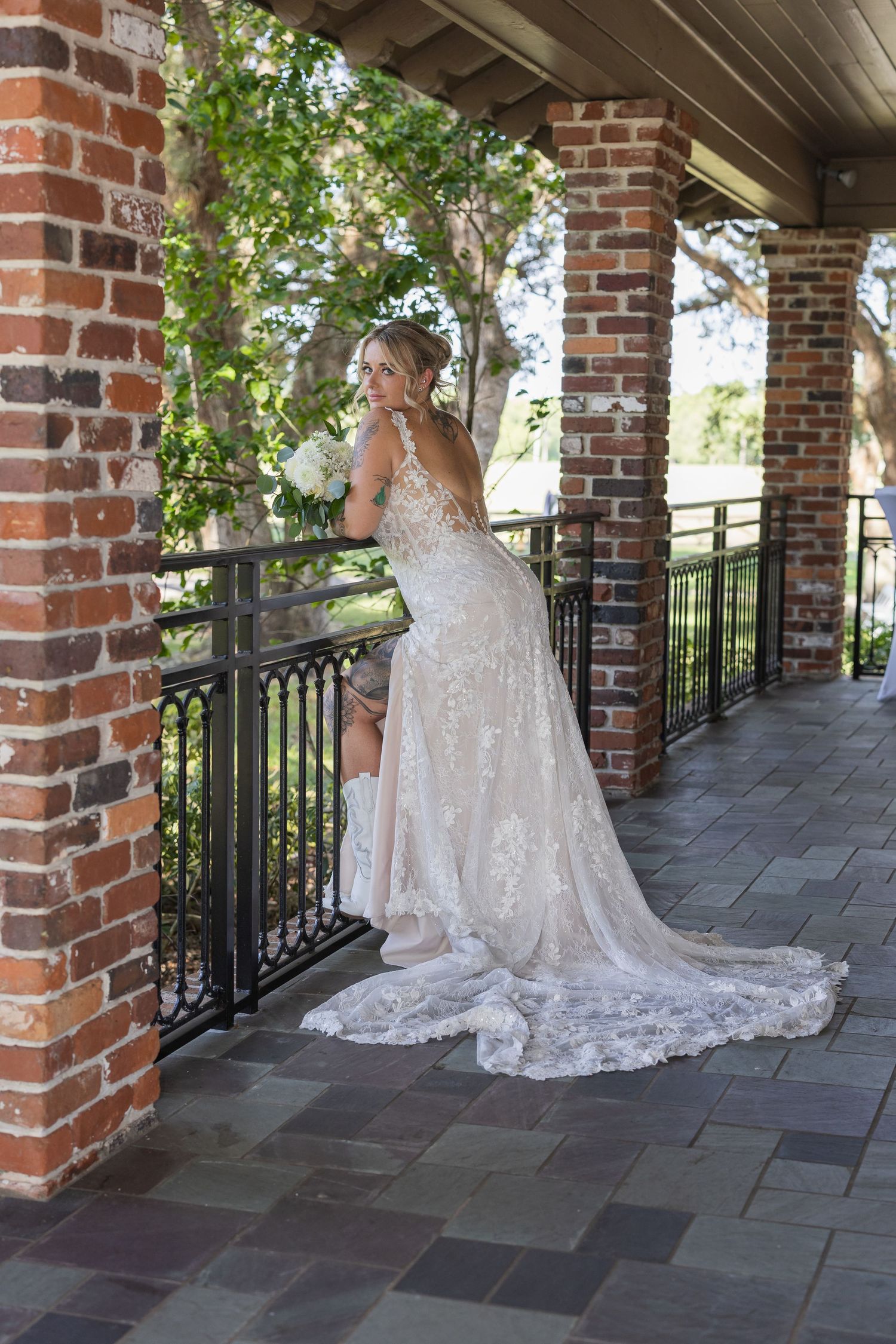 Elegant lace wedding dress with long train shown on covered brick porch with decorative railing and stone floor.