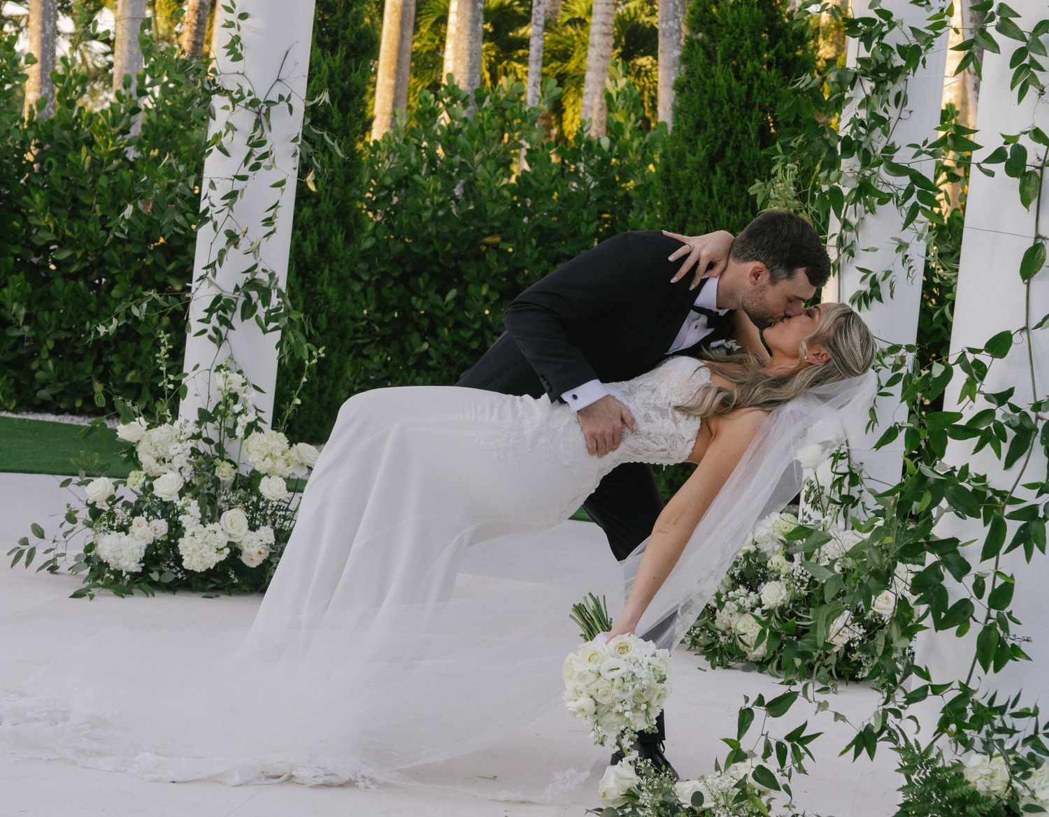 A couple in formal wedding attire dips romantically in a lush garden setting with palm trees and white floral decor.