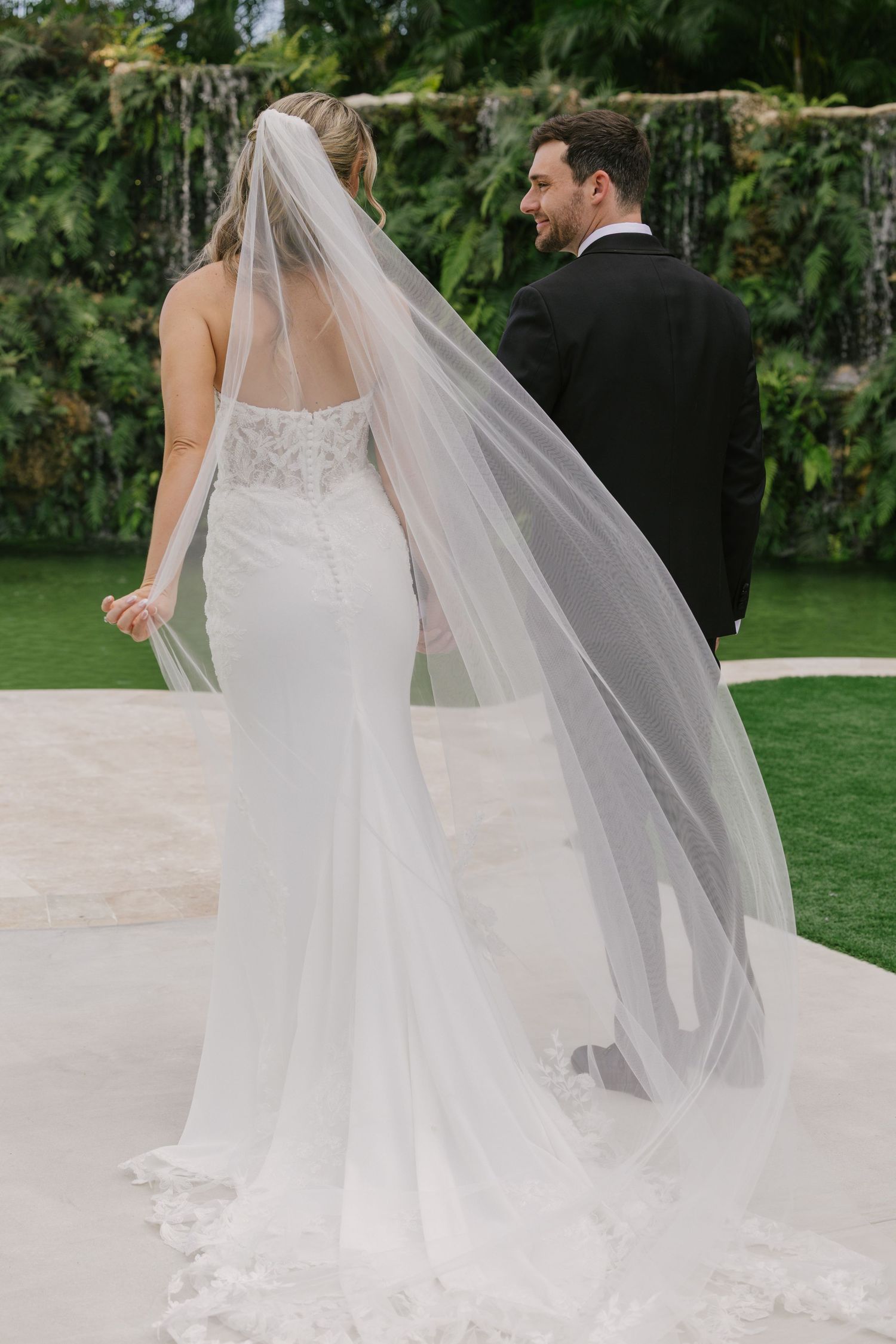 Elegant wedding couple walking outdoors with flowing white veil and lace dress details against lush garden backdrop.