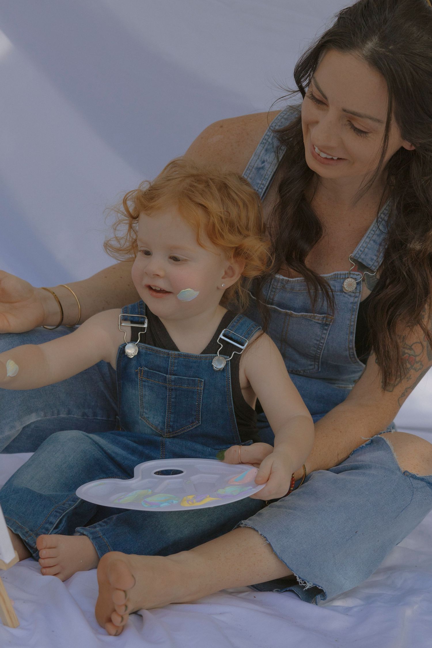 Parent and toddler in denim overalls draw together with colorful art supplies on a white surface.